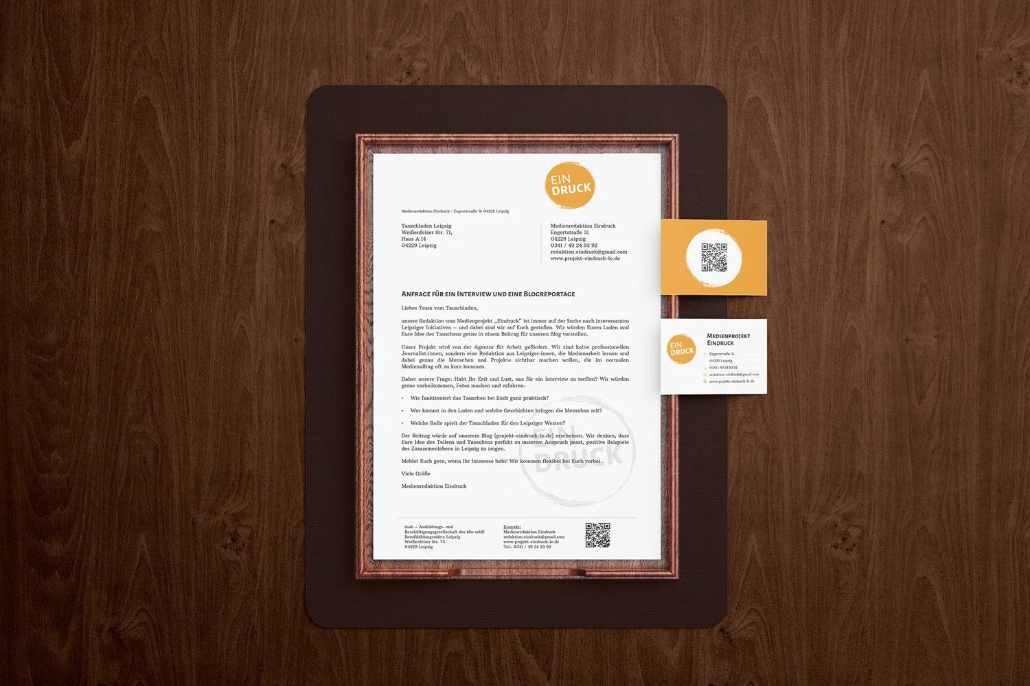 A framed letter on a desk with business cards attached. The letter contains German text and has the heading 'EIN DRUCK'. The business cards also have the same heading, one with a QR code, and include contact information.