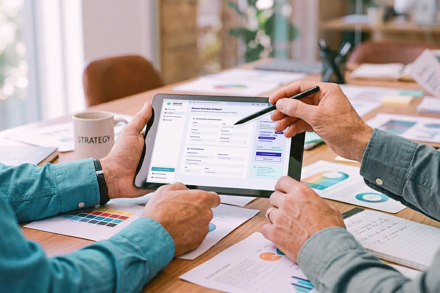 Two people reviewing data on a digital tablet at a conference table filled with papers, charts, and a coffee mug labeled 'STRATEGY'.