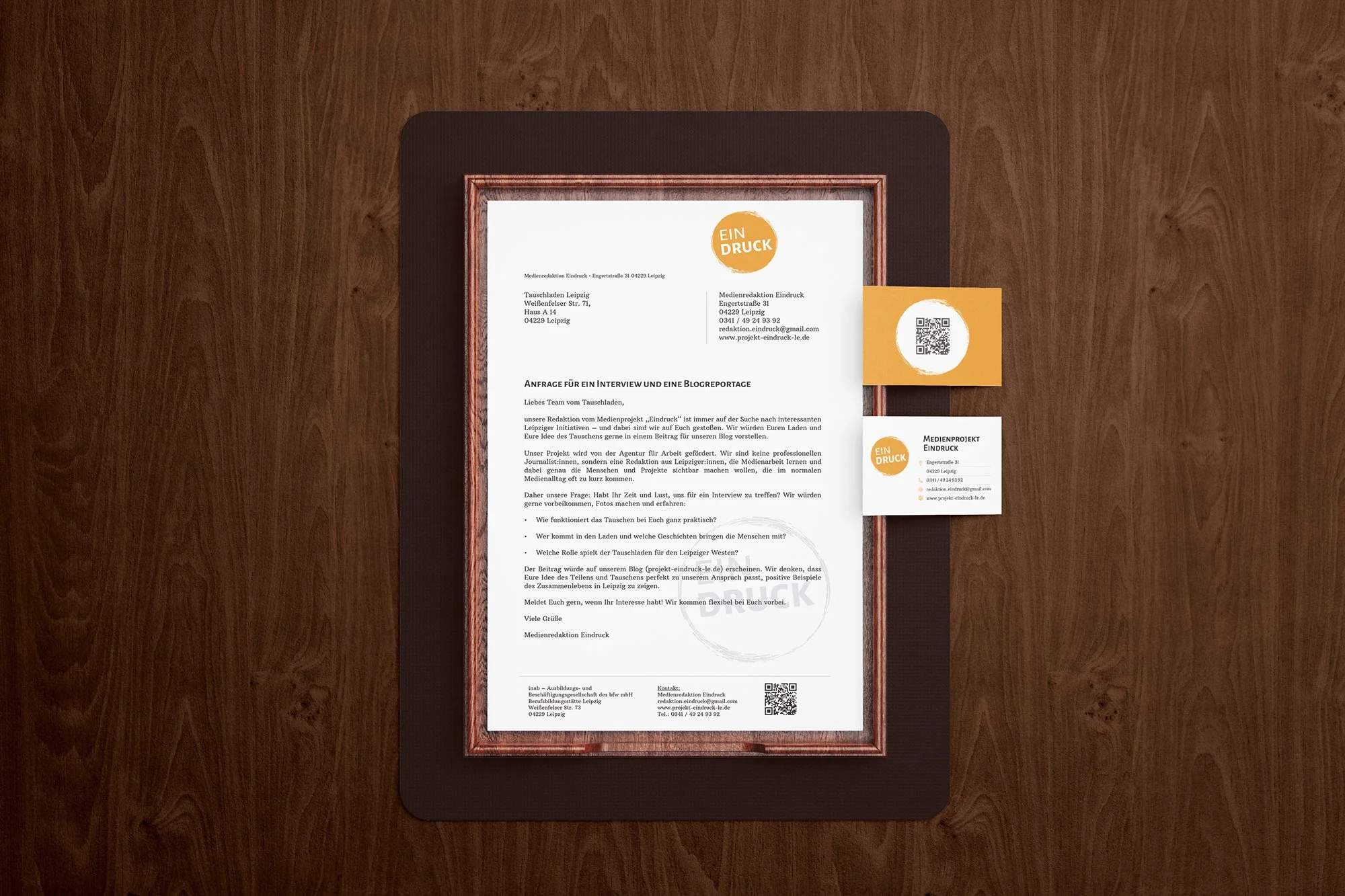 A framed document, a business card, and a QR code on a wooden surface. The document is inside a wooden frame, and the business card and QR code are positioned to the right of the frame.