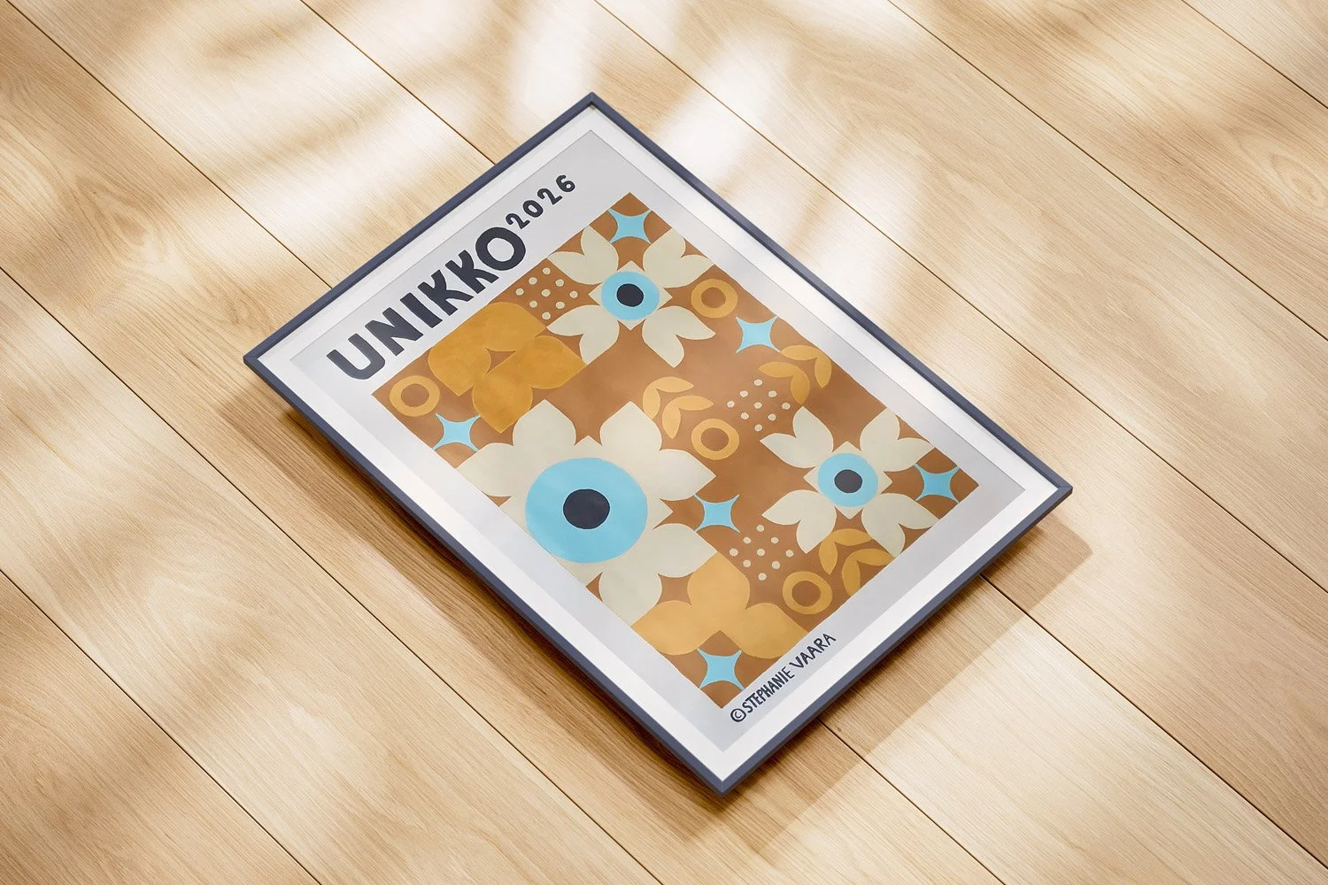 Framed art print titled 'UNIIKKO 2016' lying on a wooden floor, featuring a geometric floral pattern in shades of brown, cream, blue, and gold, with the artist's name 'Stephanie Vaar' at the bottom.