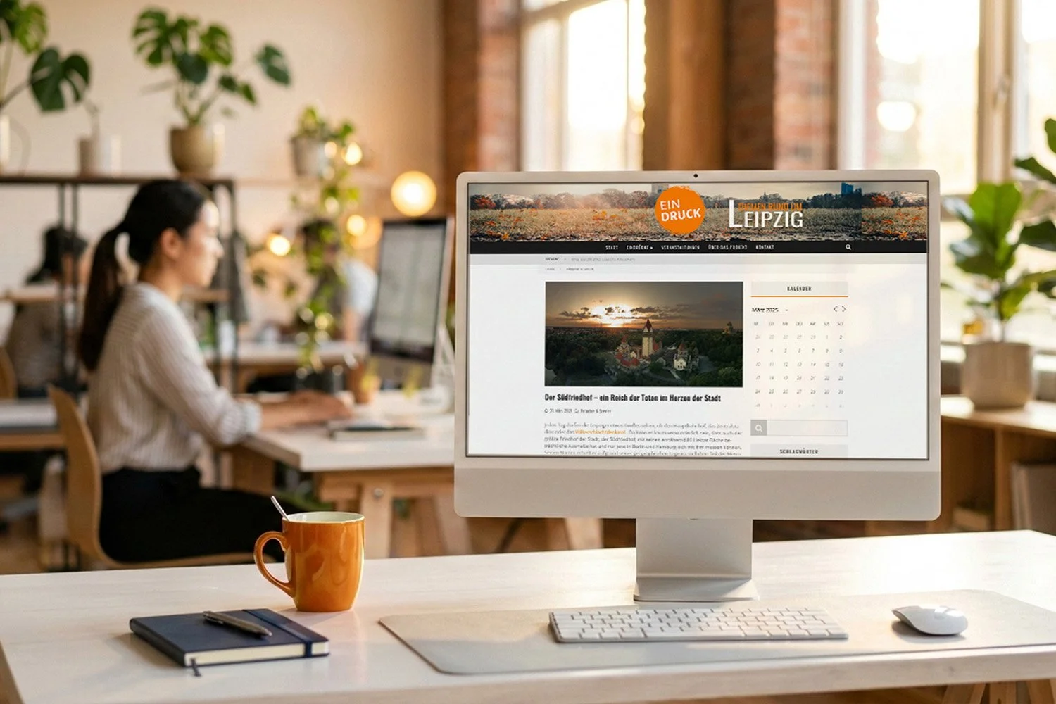 A computer monitor on a desk displays a website in German, with a photo of a sunset over a church and trees. A calendar for May 2022 is visible. The desk has a yellow mug, a closed notebook with a pen, a keyboard, and a mouse. In the background, a wo