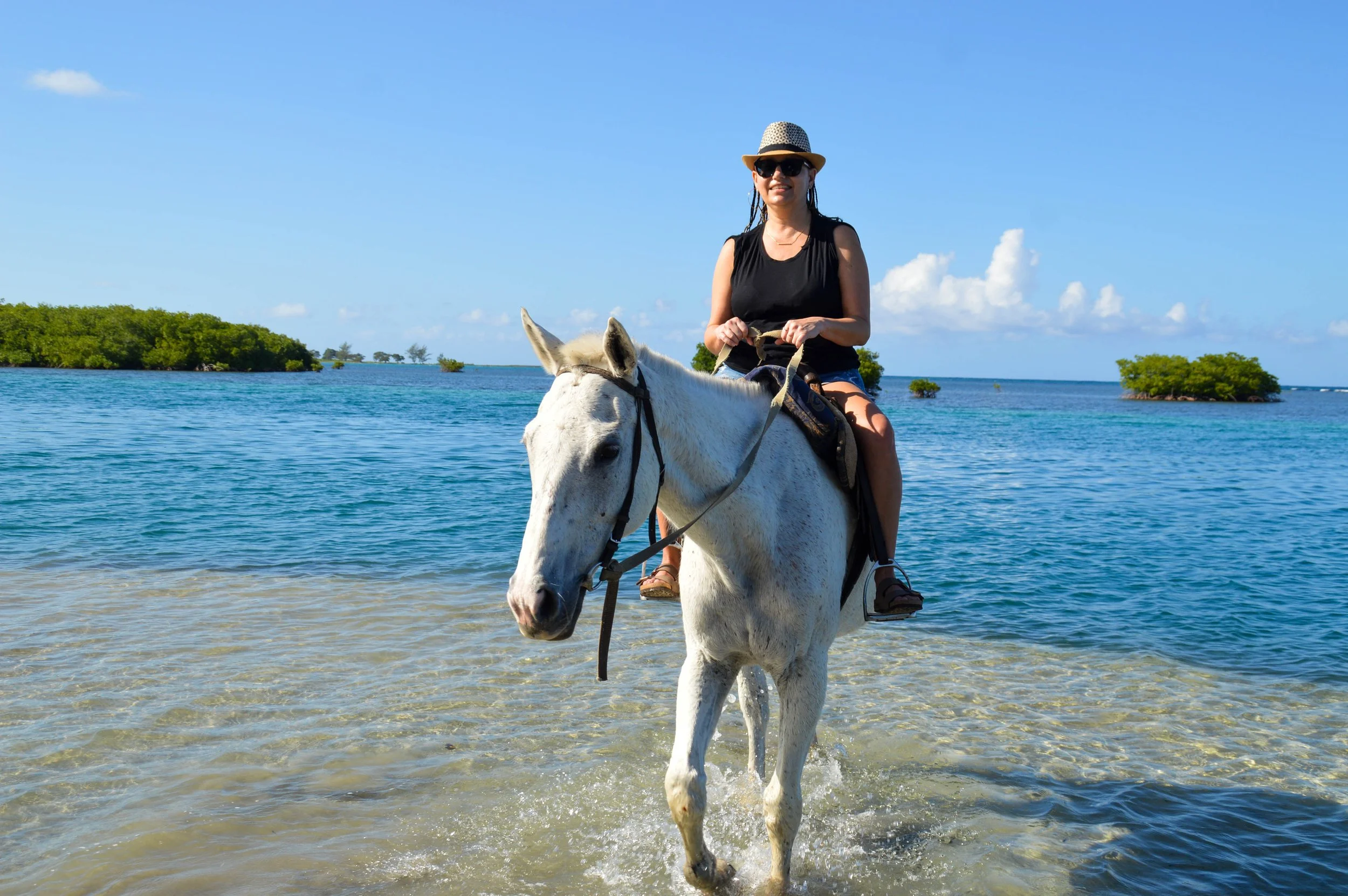Woman riding a white horse through shallow water at the beach with small islands and blue sky in the background.
