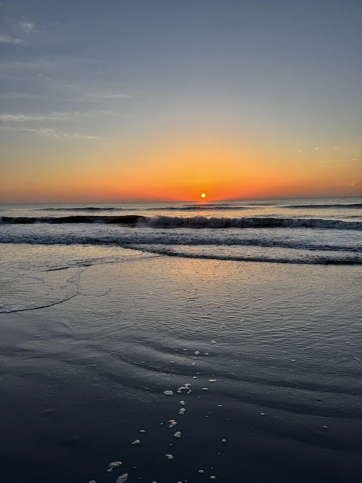 Sunset over the ocean with gentle waves and foam on the water, reflecting a colorful sky with hues of orange, pink, and blue.