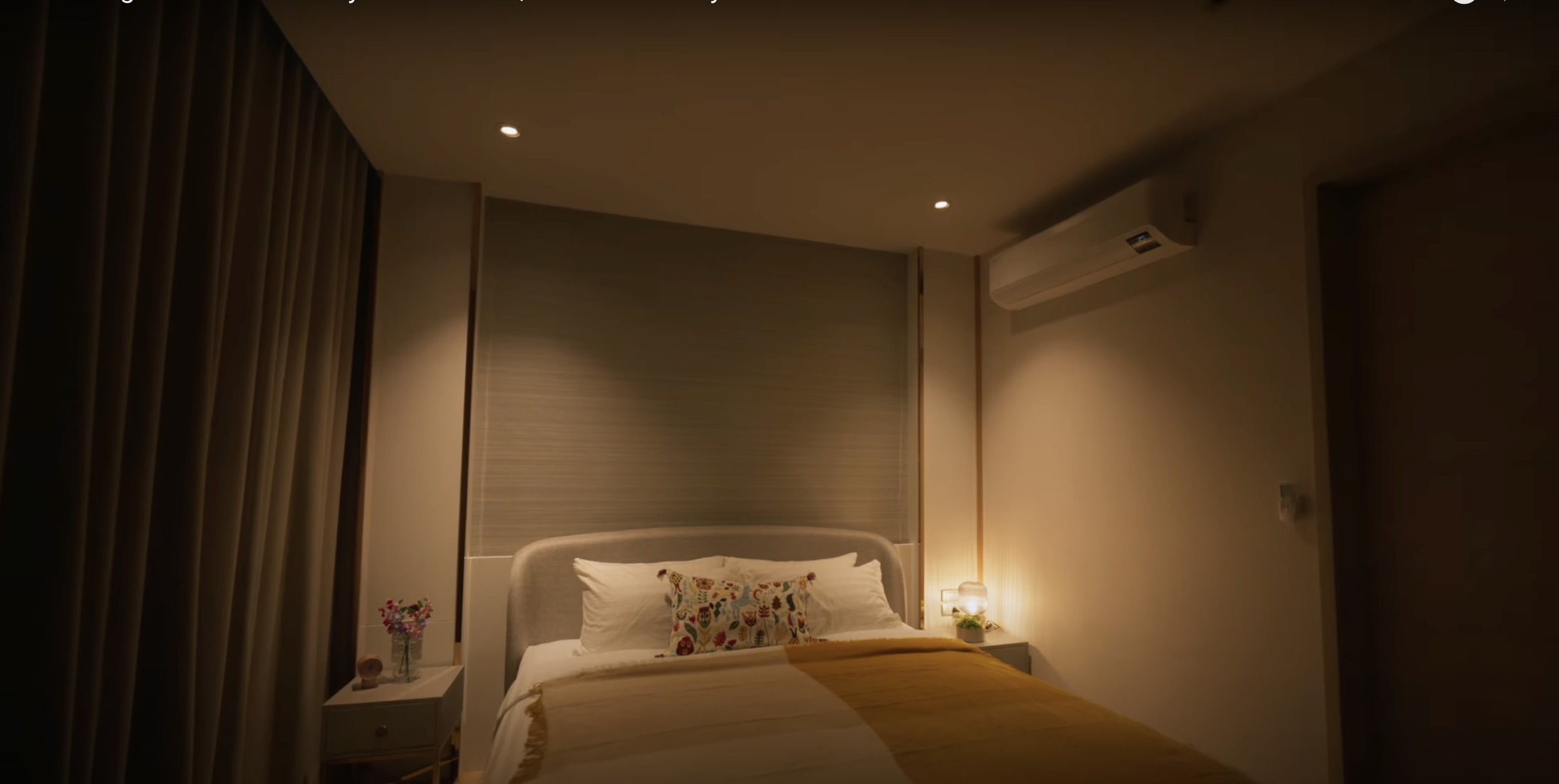 A warmly lit hotel bedroom with a bed, pillows, bedside tables, a small flower arrangement, and modern decor.