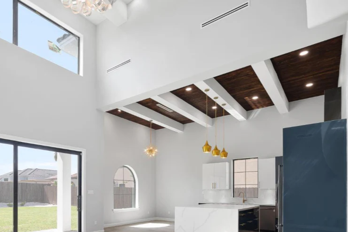 Modern kitchen with high vaulted ceiling, dark wood beams, white walls, large windows, and a central island with white marble countertop.