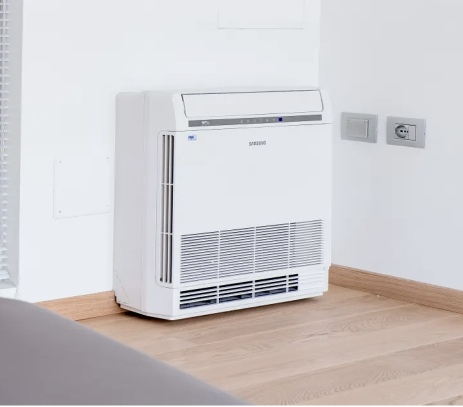 A Samsung portable air conditioner unit placed on a wooden floor near a white wall and electrical outlets.