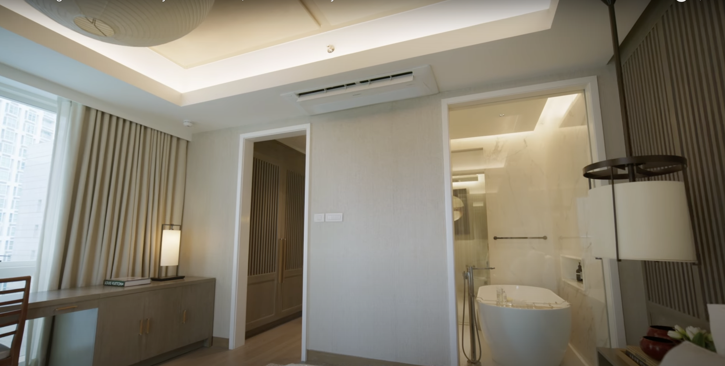 Modern hotel room with a bathroom featuring a bathtub and marble walls, a desk with a lamp, and floor-to-ceiling curtains by the window.