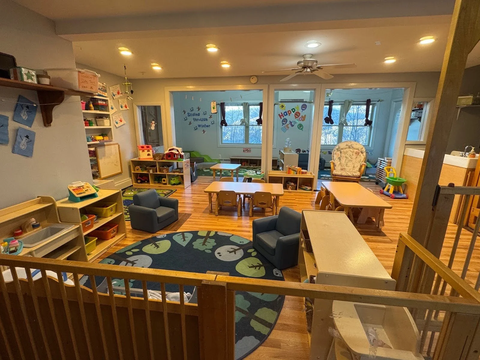 A colorful classroom with play tables, chairs, shelves filled with toys, a circular rug with a tree pattern, and a cozy reading nook with windows and holiday decorations.