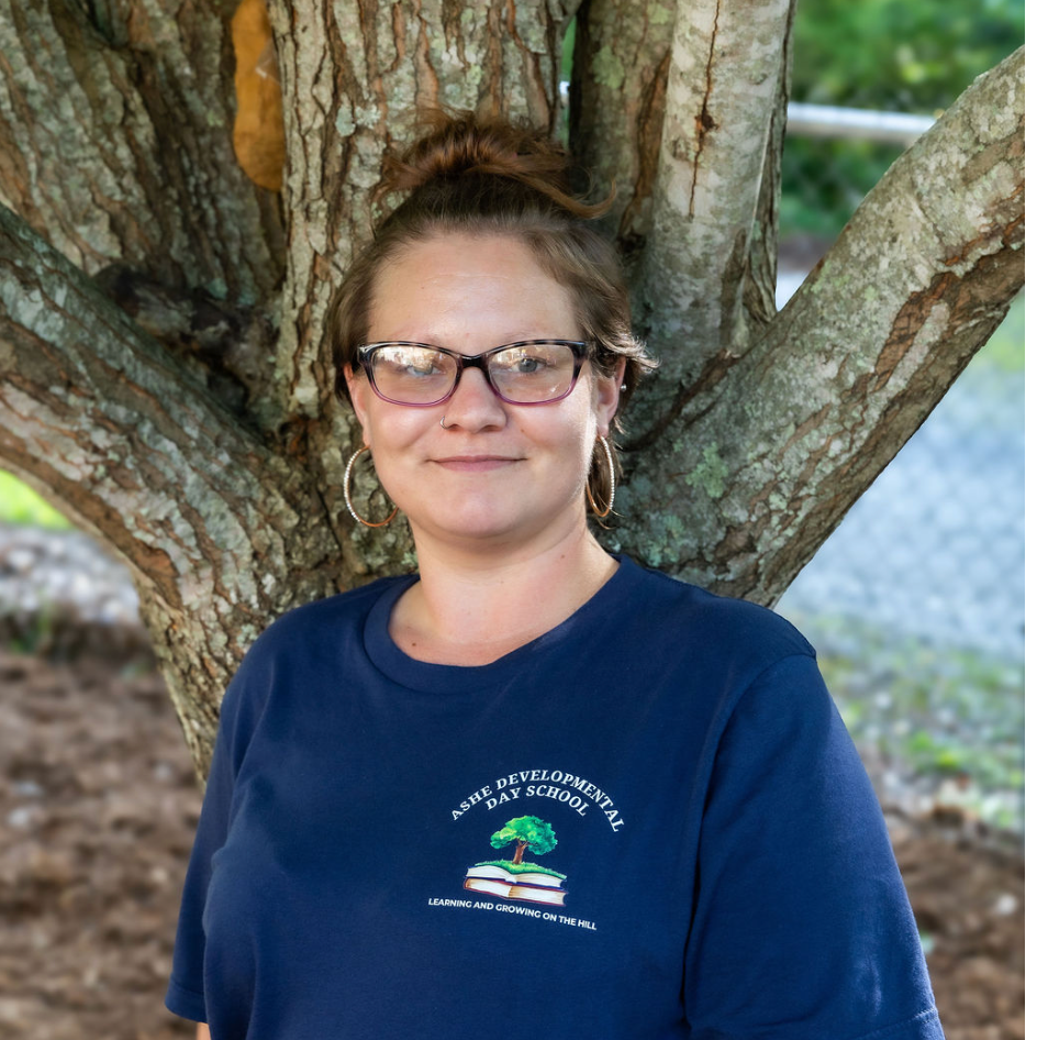 A woman with glasses, hoop earrings, and a bun hairstyle, standing outdoors in front of a tree with textured bark.