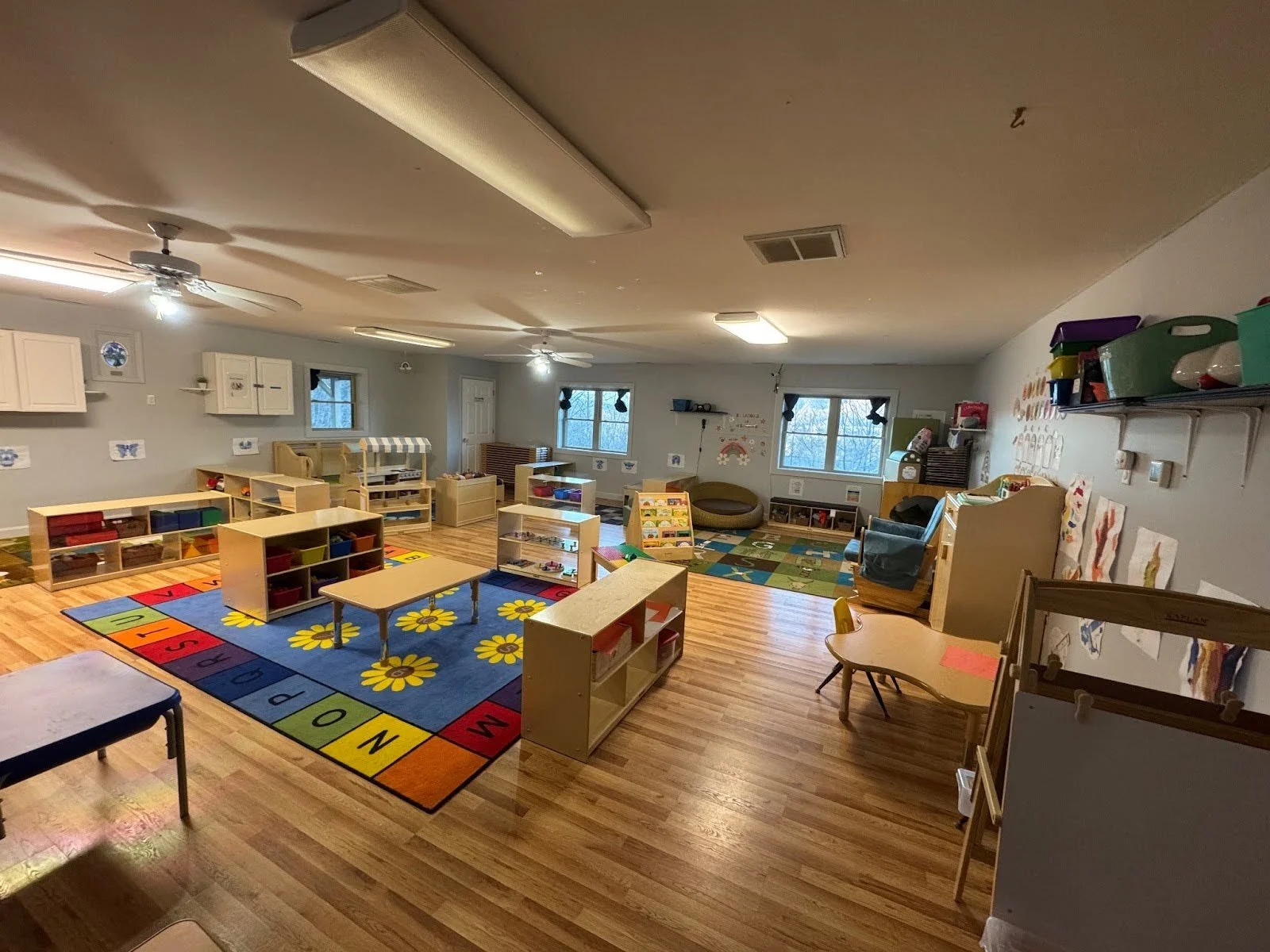 Empty preschool classroom with colorful area rugs, shelves filled with toys and books, small tables and chairs, and windows letting in natural light.