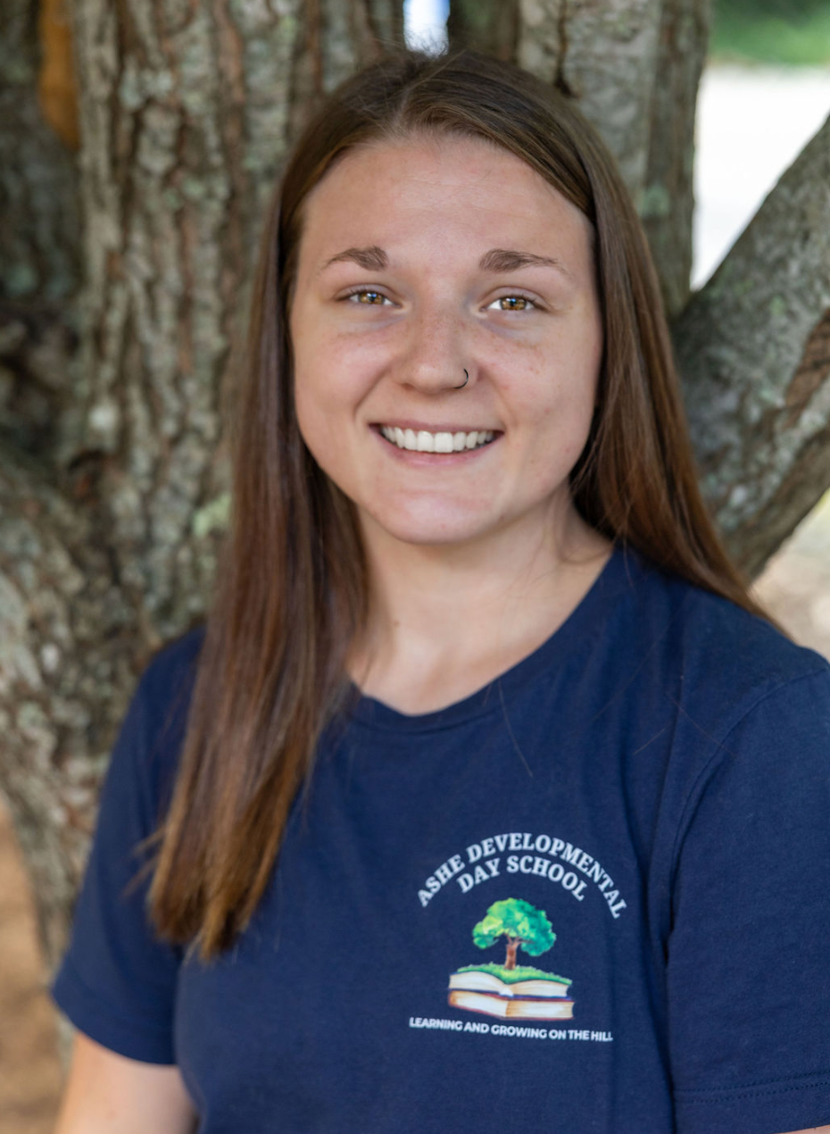 A young woman with long brown hair, light skin, and a nose piercing, smiling, standing outdoors in front of a tree, wearing a navy blue T-shirt with a logo that reads "Ashé Developmental Day School" and a picture of a tree, a book, and the phrase "Learning and Growing on the Hill."
