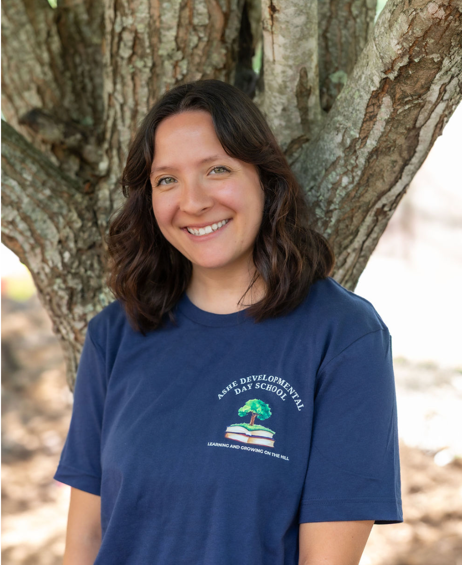 A woman with shoulder-length brown hair smiling, wearing a navy blue T-shirt with a logo and text, standing in front of a large tree trunk outdoors.