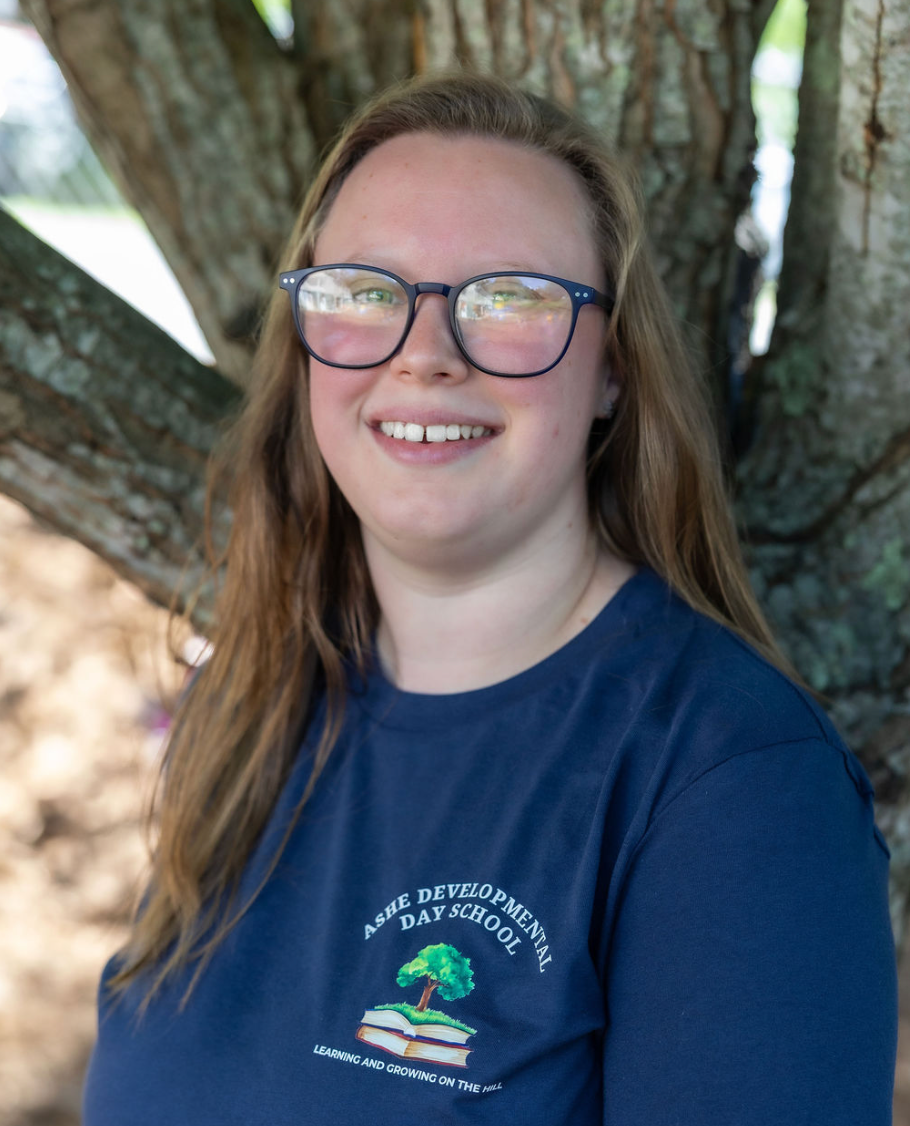 A young woman with long, light brown hair, wearing glasses and a navy blue t-shirt with the logo for Ashe Developmental Day School, smiling outdoors with a large tree in the background.