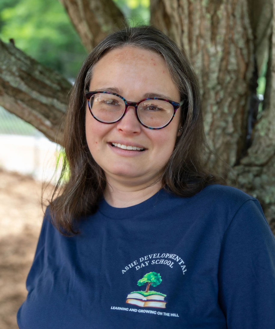A woman with long brown hair and glasses is smiling outdoors in front of a large tree. She is wearing a navy blue t-shirt with a logo that reads "ASHE DEVELOPMENTAL DAY SCHOOL" and features a tree growing out of an open book.