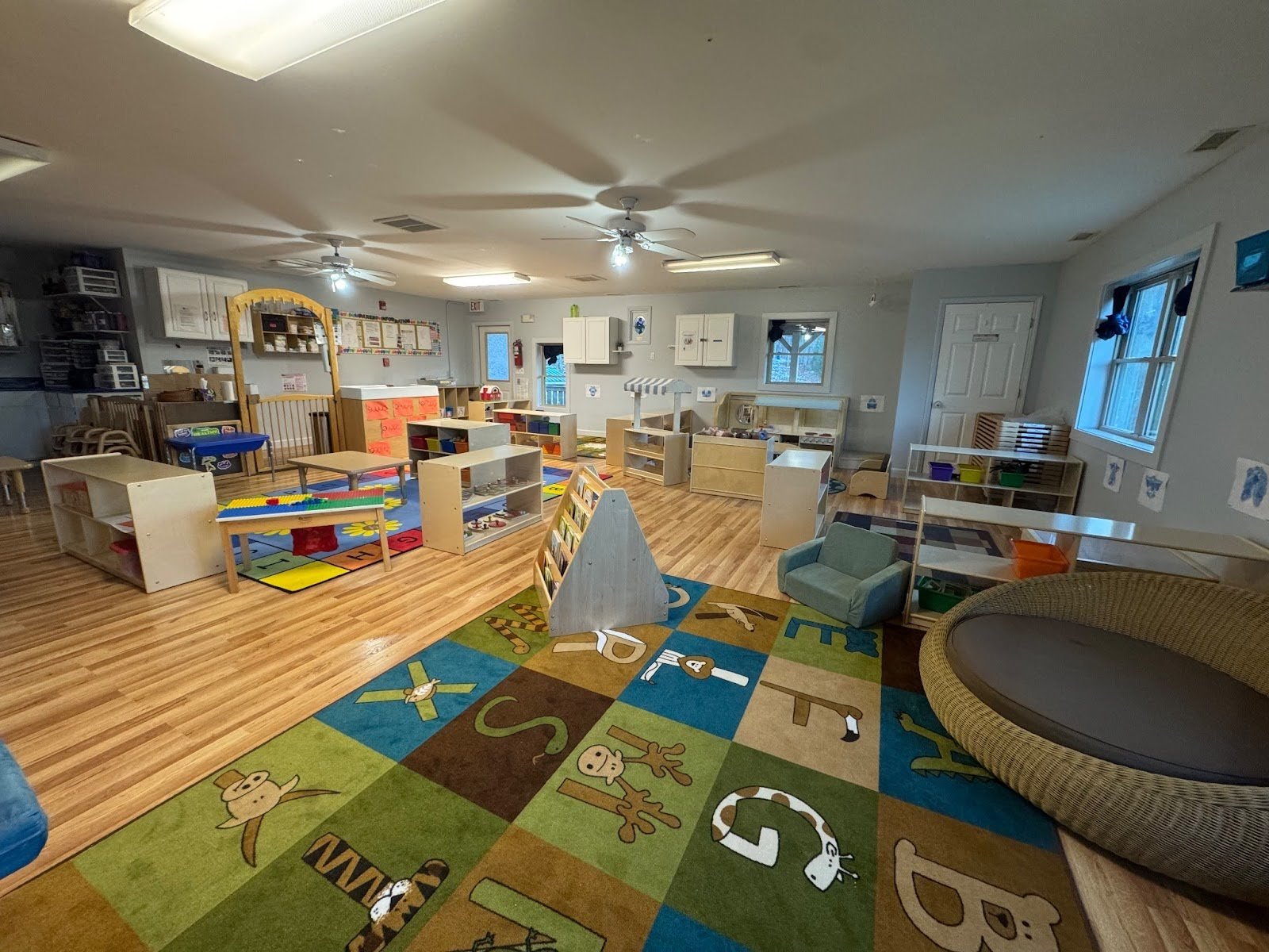 Empty preschool classroom with colorful alphabet-themed rug, wooden furniture, and educational toys scattered around.