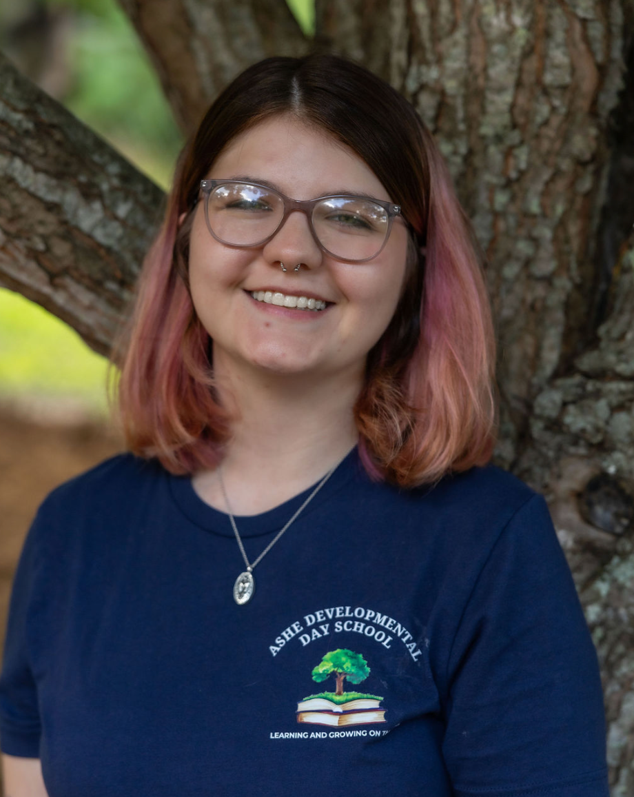 A young woman with glasses and a septum piercing smiling while standing outdoors in front of a large tree, wearing a navy blue shirt with a school logo that says "ASHE DEVELOPMENTAL DAY SCHOOL."
