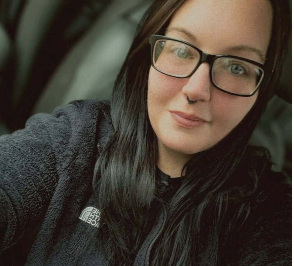 A woman with long black hair and glasses, wearing a black fleece jacket, taking a selfie indoors.