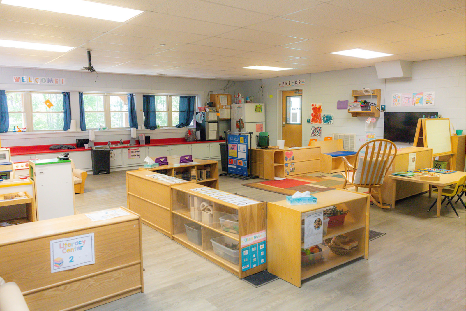 A colorful preschool classroom with various activity areas, shelves filled with toys and books, a small rug, a wooden rocking chair, and windows covered with blue curtains.