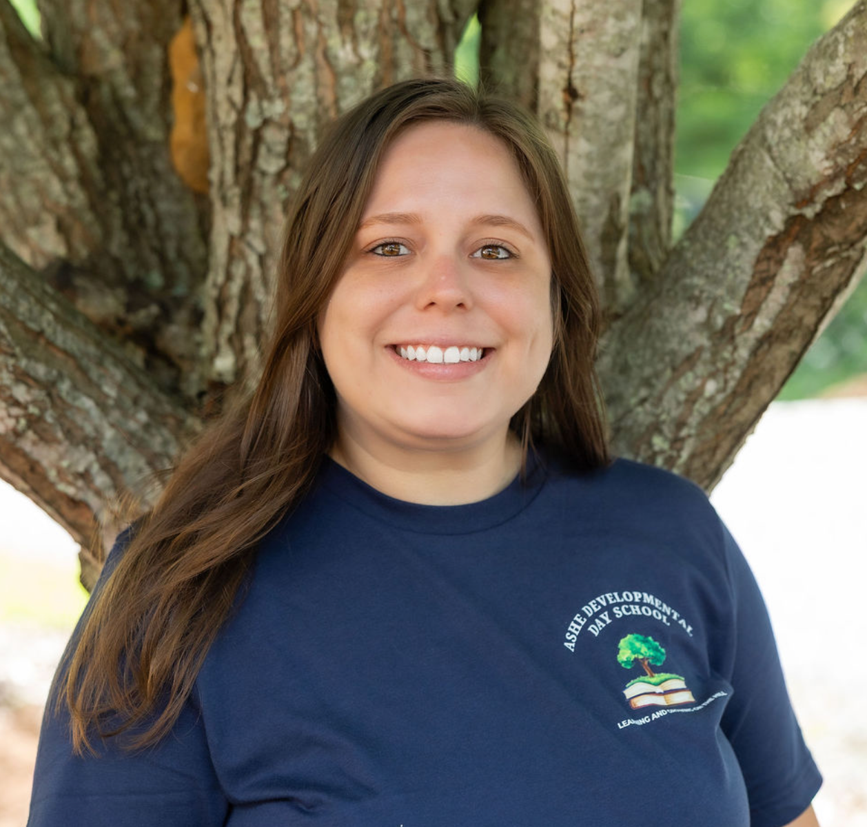 A woman with long brown hair hair standing outdoors near a tree with a background of greenery and sunlight.