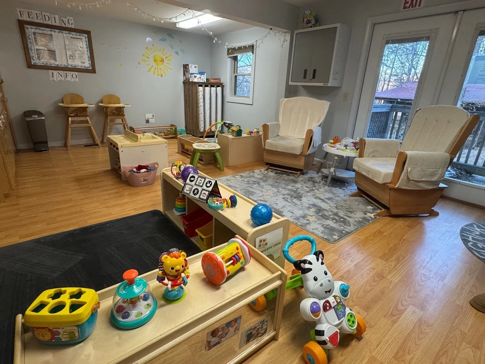 Room with children's toys and seating area, including a zebra ride-on toy, two beige armchairs, and a small play table, with large windows and a wooden floor.