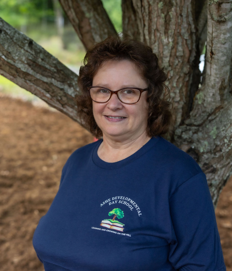 A woman with curly brown hair, wearing glasses and a navy blue T-shirt with the logo for Ashe Developmental Day School, standing outdoors in front of a large tree.