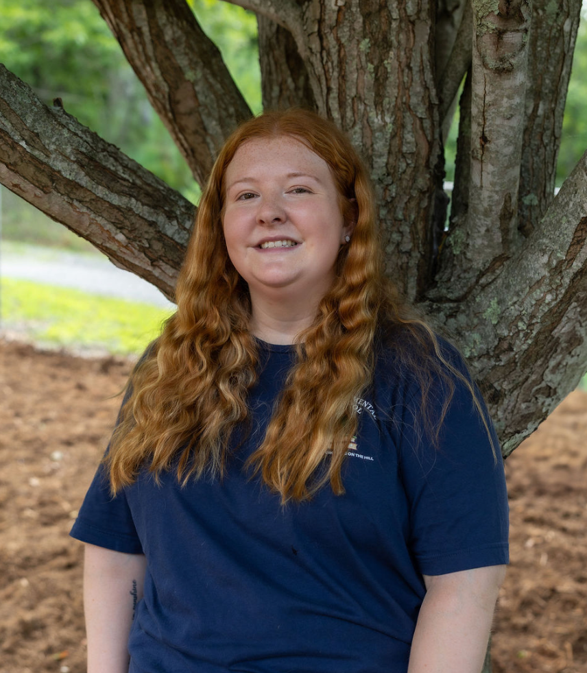 A young woman with long, curly red hair smiling and standing outdoors in front of a large tree with textured bark. She is wearing a navy blue T-shirt and has a natural background of green trees and a dirt patch.