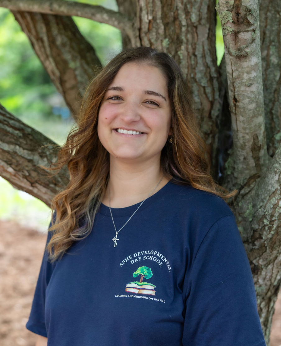 Young woman with long, wavy brown hair smiling, standing outdoors beside a large tree, wearing a navy blue t-shirt with the logo for Ashe Developmental Day School.