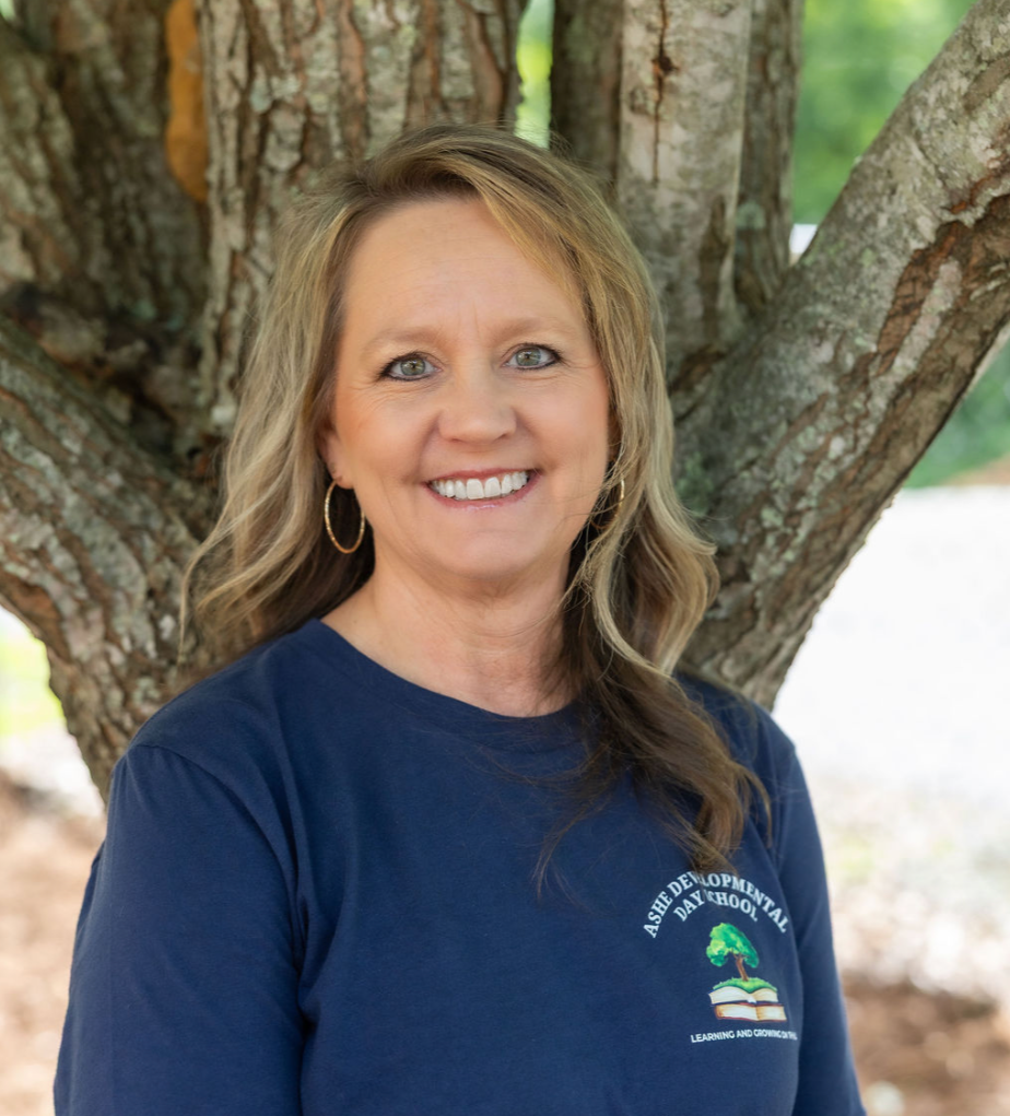 A woman with light brown hair, hoop earrings, and a blue shirt with the logo 'Ashe Developmental Day School' stands outdoors in front of a tree, smiling.