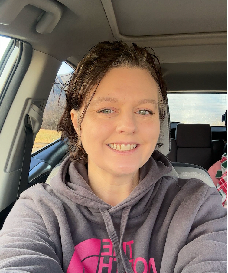 A woman with short, dark, curly hair smiling inside a vehicle. She is wearing a gray hoodie with pink letters on it. The background shows the vehicle's interior and a glimpse of the outside scenery through the windows.