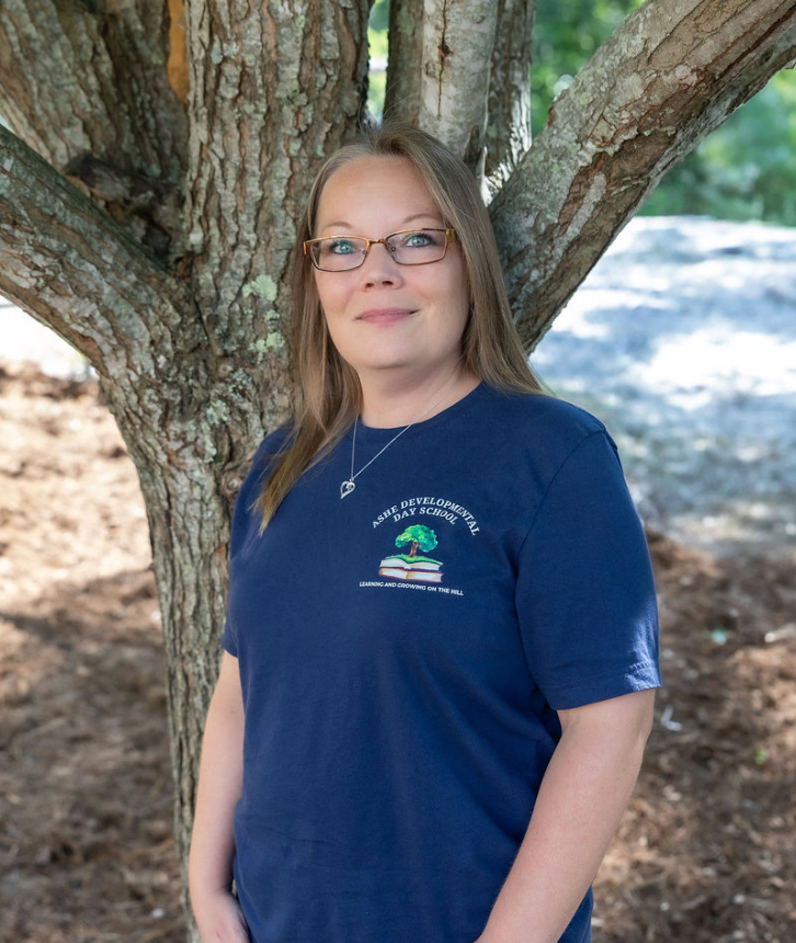 A woman with short curly red hair and glasses standing outdoors in front of a tree, wearing a navy blue t-shirt with a logo of a tree and books that reads 'Ashe Developmental Day School'.