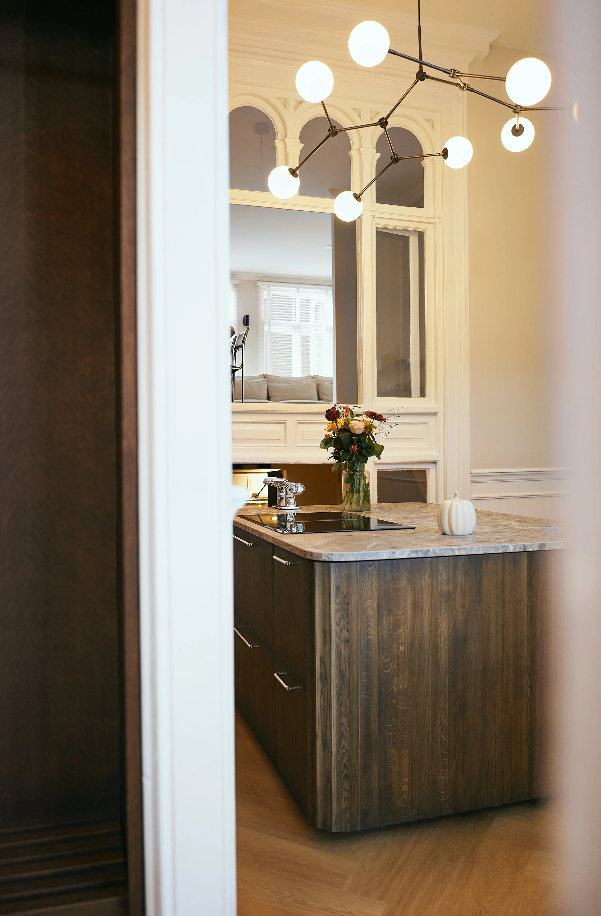 View of a custom build curved kitchen island with a marble countertop, dark wood cabinets and modern light fixtures above. Foreground is a walnut custom wardrobe Background includes a mirror and a living room with sofas and large windows.