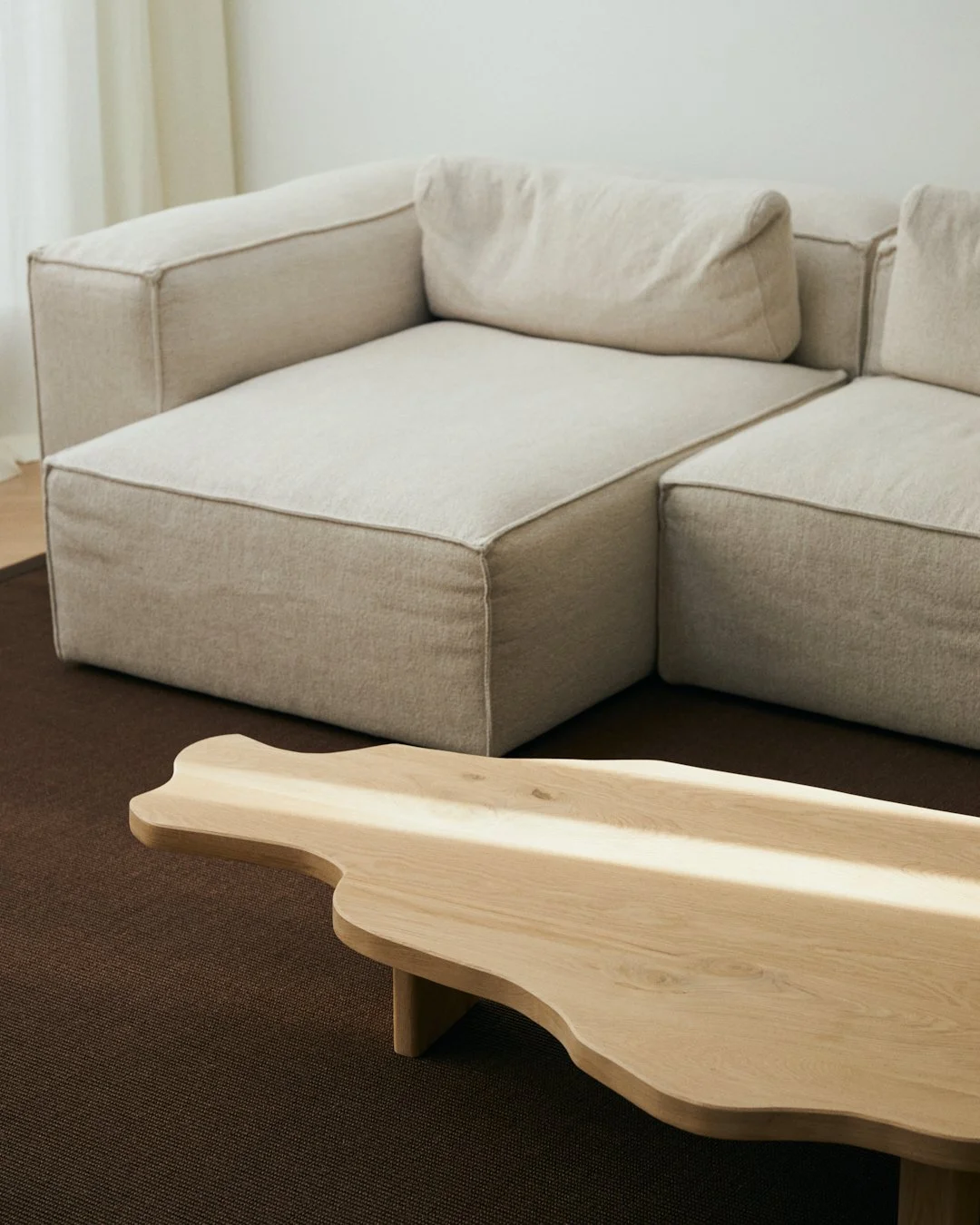 A beige sectional sofa with cushion back rests, positioned next to a wooden bespoke coffee table with a natural shape, on a dark brown carpet.