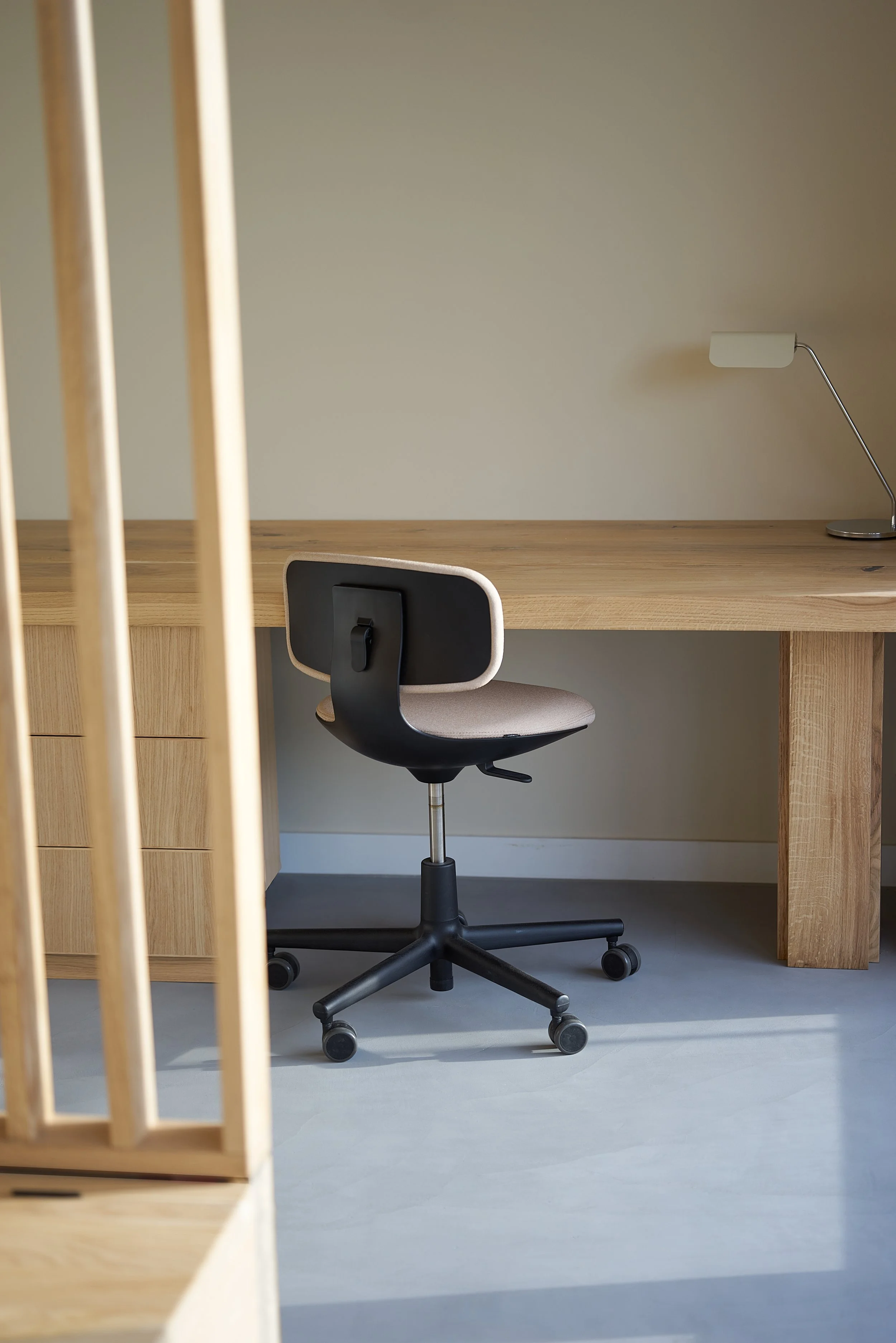 An japanese modern office desk with a black and beige swivel chair, a white desk lamp, and a wooden side cabinet against a plain wall. in the foreground a solid oak roomdivider and light grey micro-cement floors