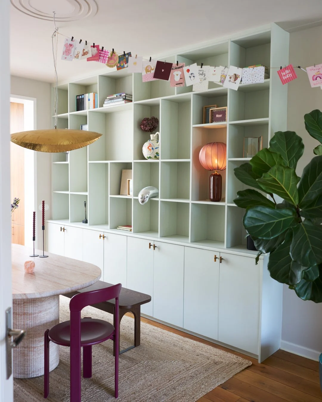 Living room with a  custom made shelving unit decorated with books, vases, and a paper lantern, a green plant, a wooden dining table, a purple chair, and colorful hanging greeting cards.