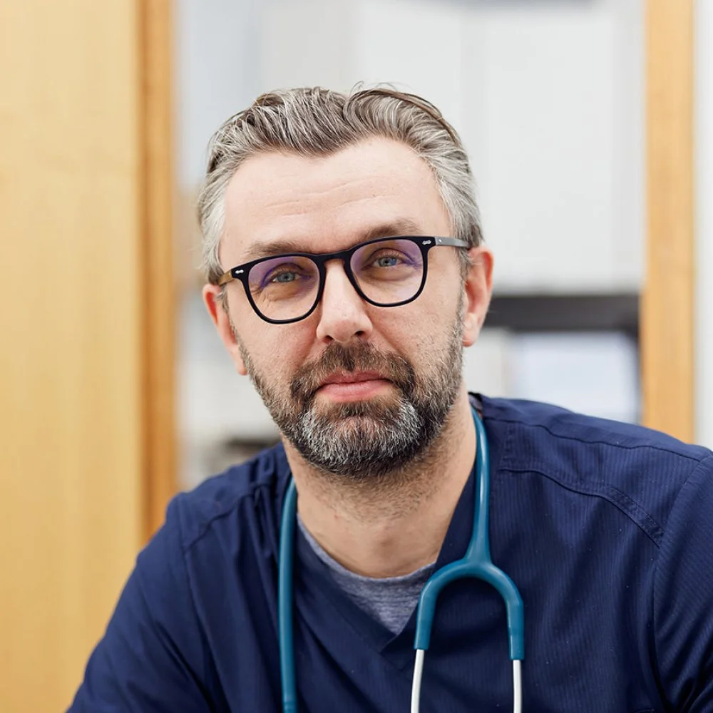 A man wearing glasses and a stethoscope around his neck, dressed in scrubs, looking at the camera in a clinical setting.