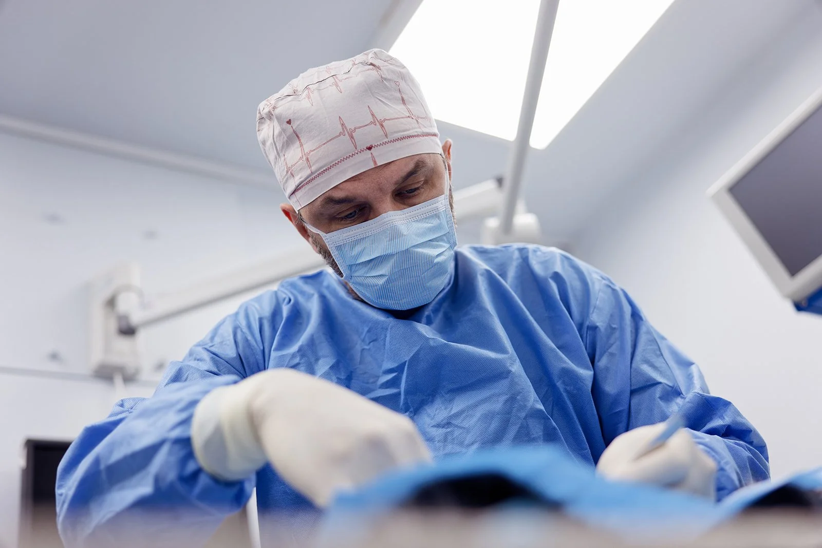 A male surgeon in scrubs, surgical mask, and cap performing a procedure in an operating room.