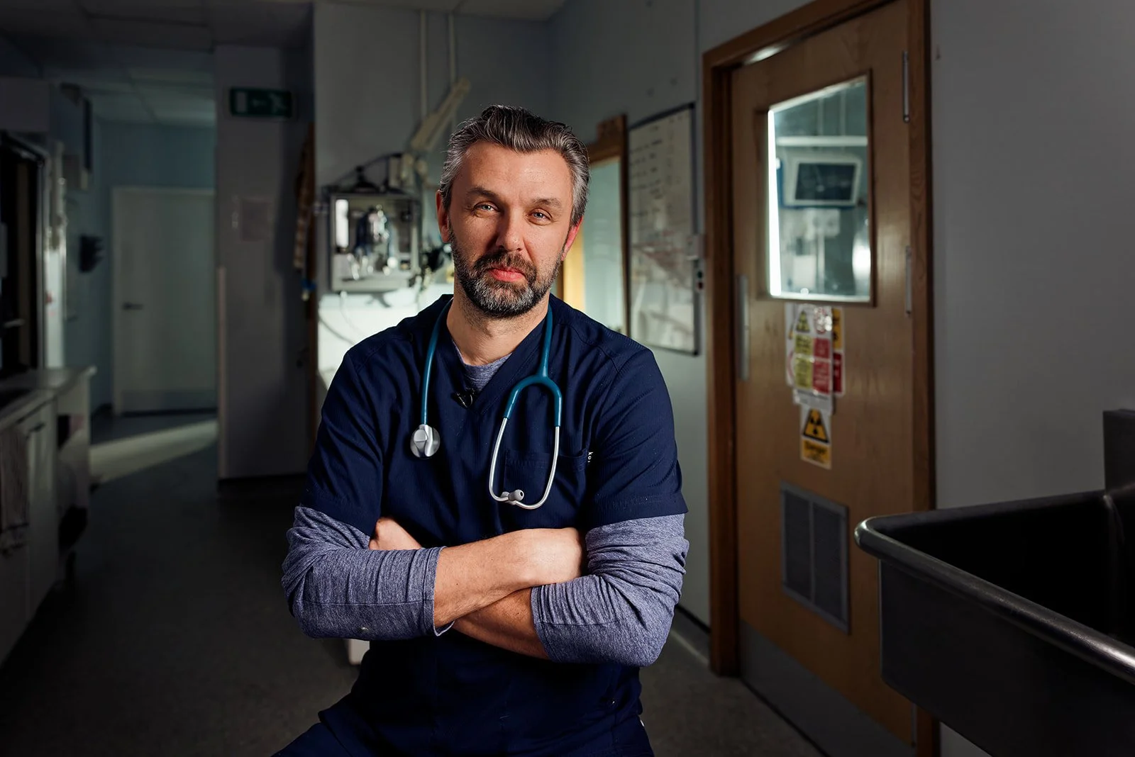 A male healthcare professional with a beard and gray hair, wearing dark scrubs and a stethoscope, standing in a hospital or clinic corridor with arms crossed.