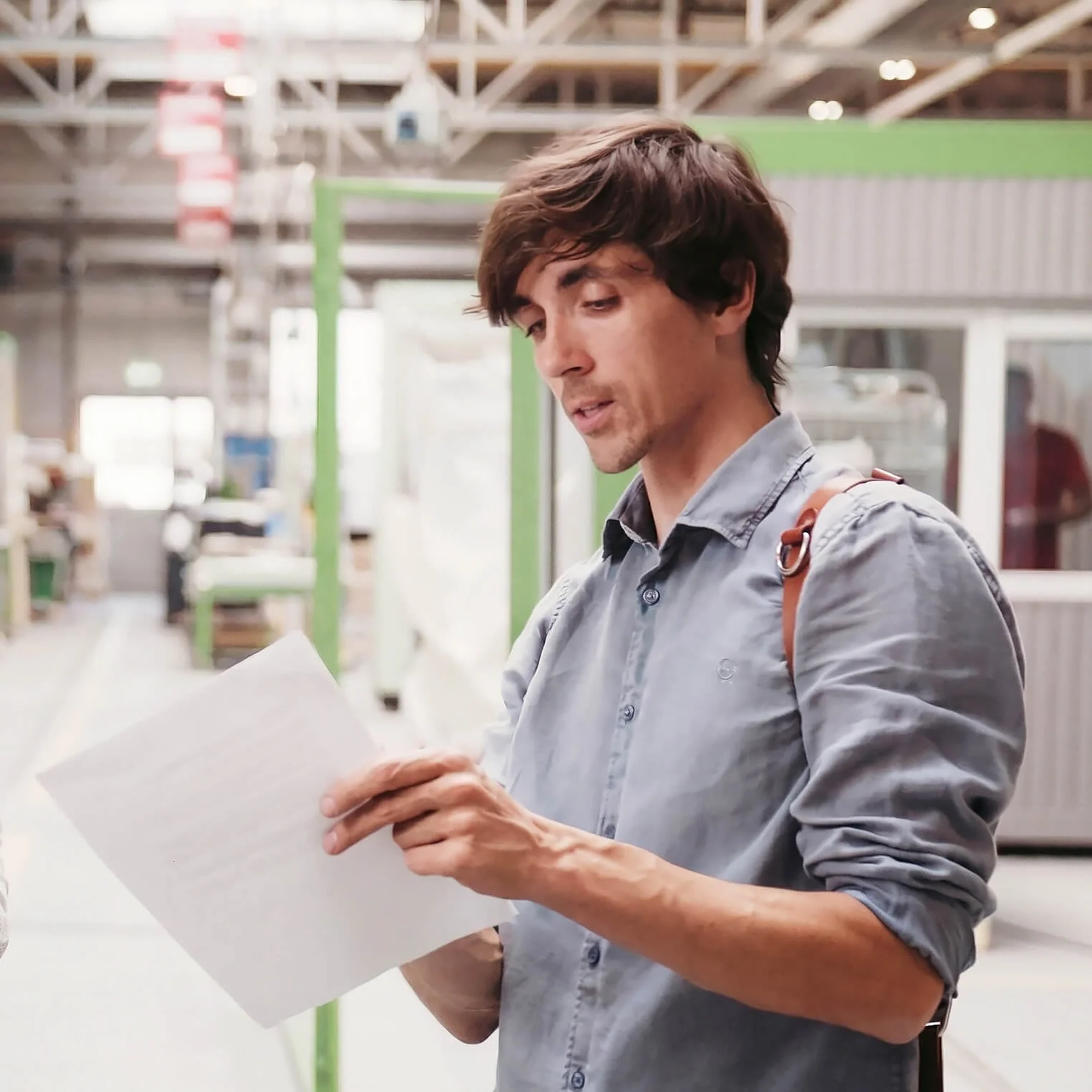 A man with dark hair wearing a gray shirt is reading a piece of paper inside a store or warehouse.
