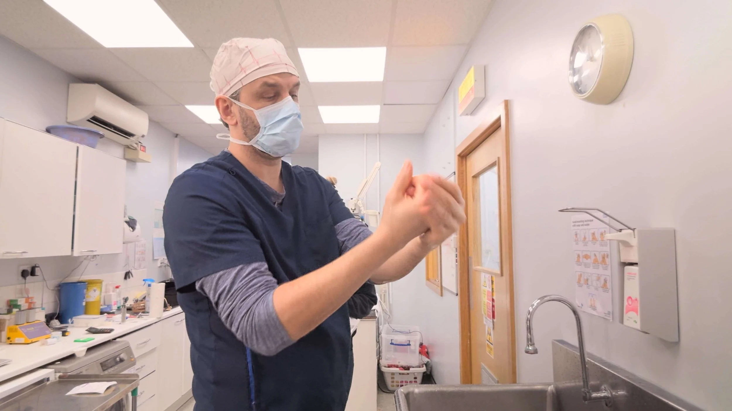 A healthcare professional wearing scrubs, a face mask, and a surgical cap, standing in a medical facility, preparing a medical procedure or administering treatment.