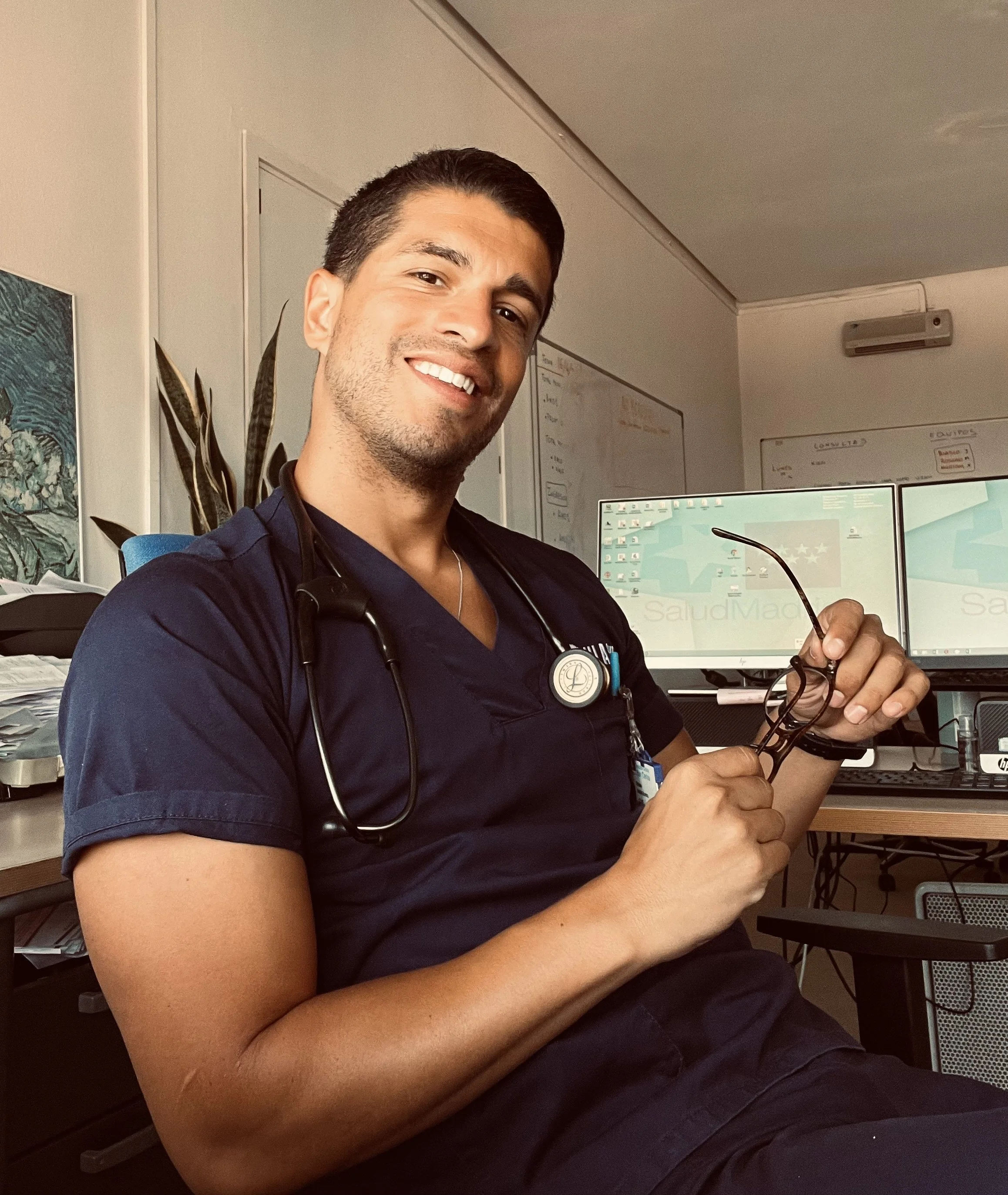 Un hombre joven, médico, con uniforme de enfermería y estetoscopio, sentado en una oficina, sosteniendo sus gafas, sonriendo a la cámara.