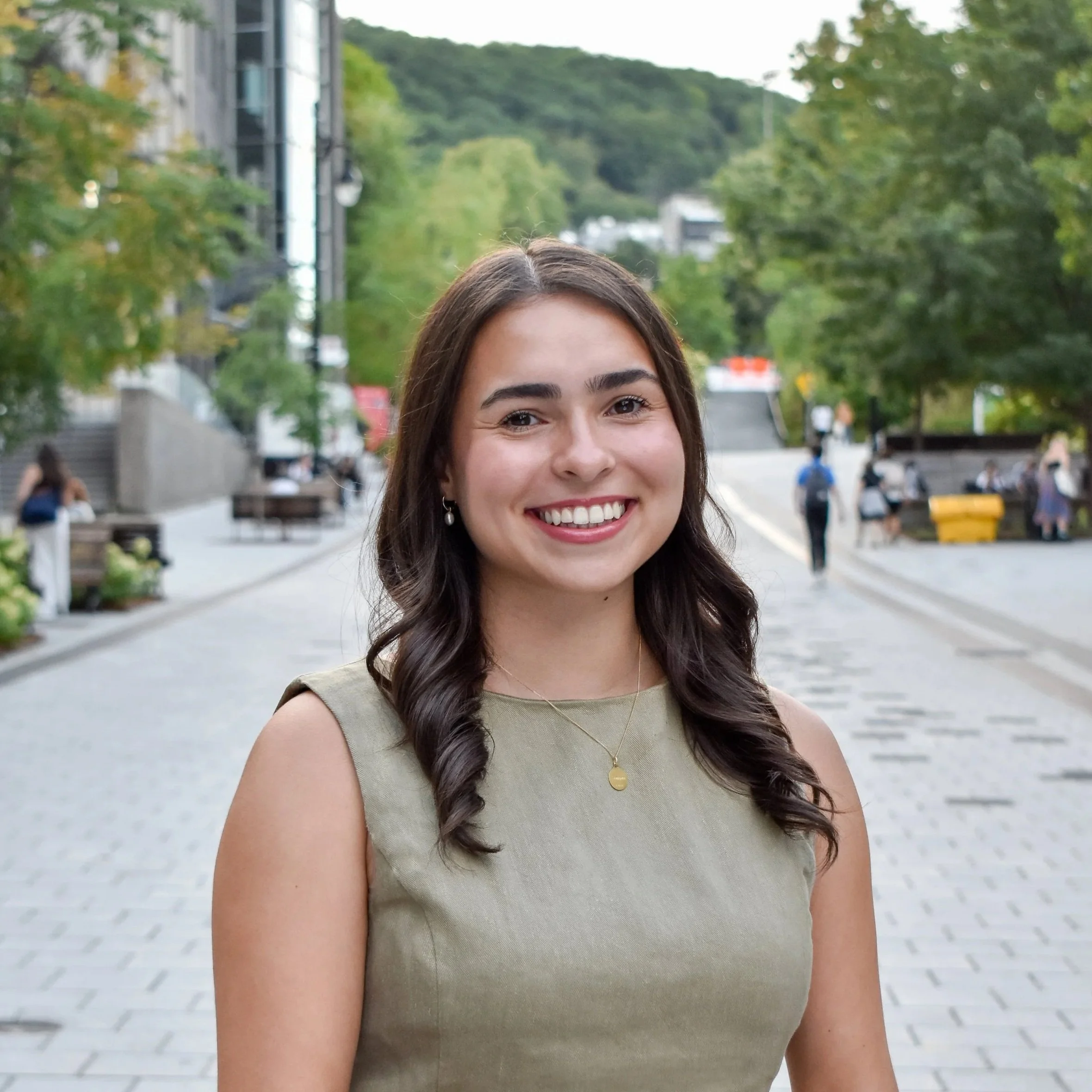 A young woman with dark brown hair styled in loose curls, smiling outdoors on a city street with trees, benches, and other people in the background.