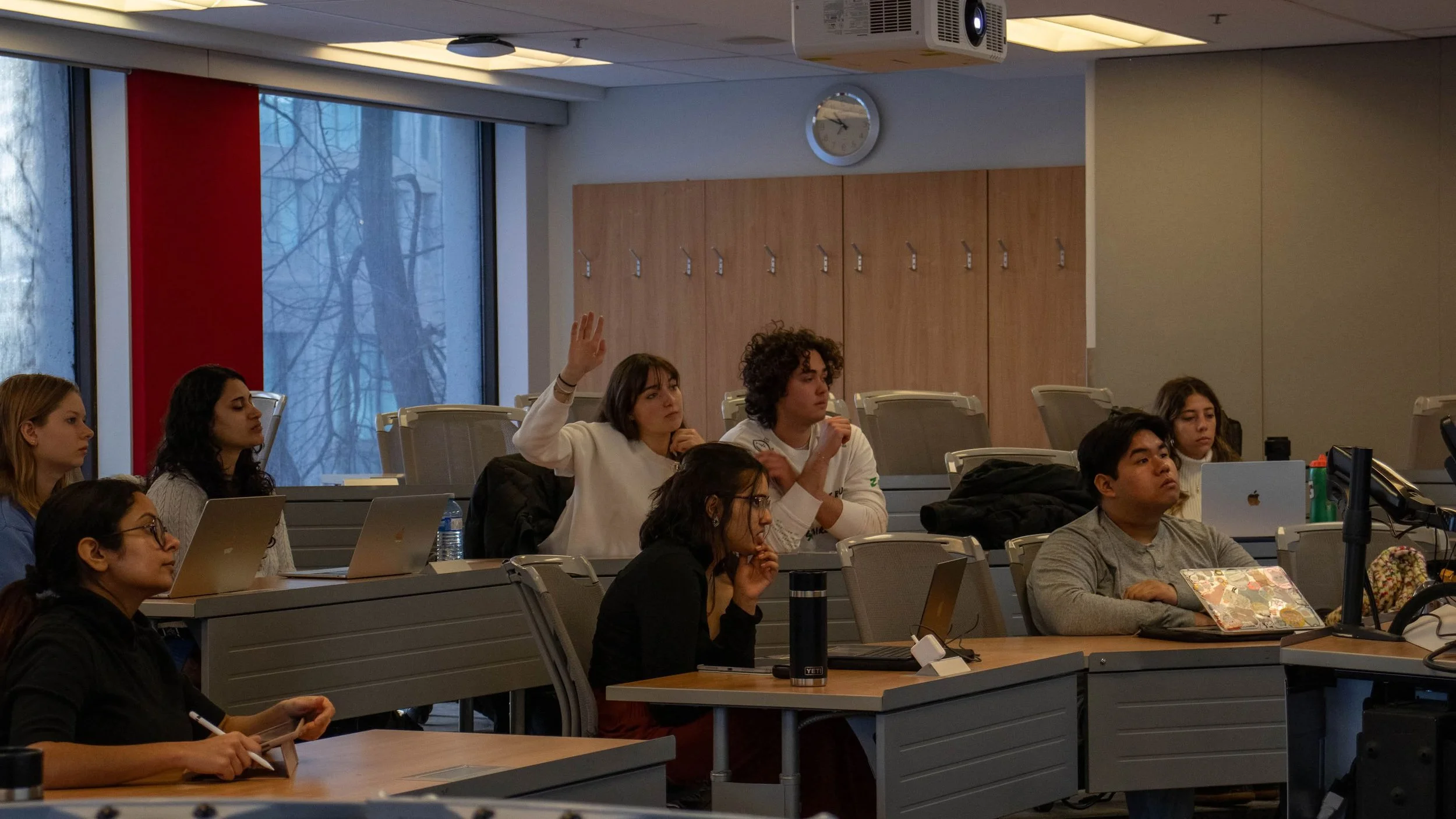 Students attending a lecture in a classroom, with some raising their hands, sitting at desks with laptops and notebooks, and a large window showing trees outside.