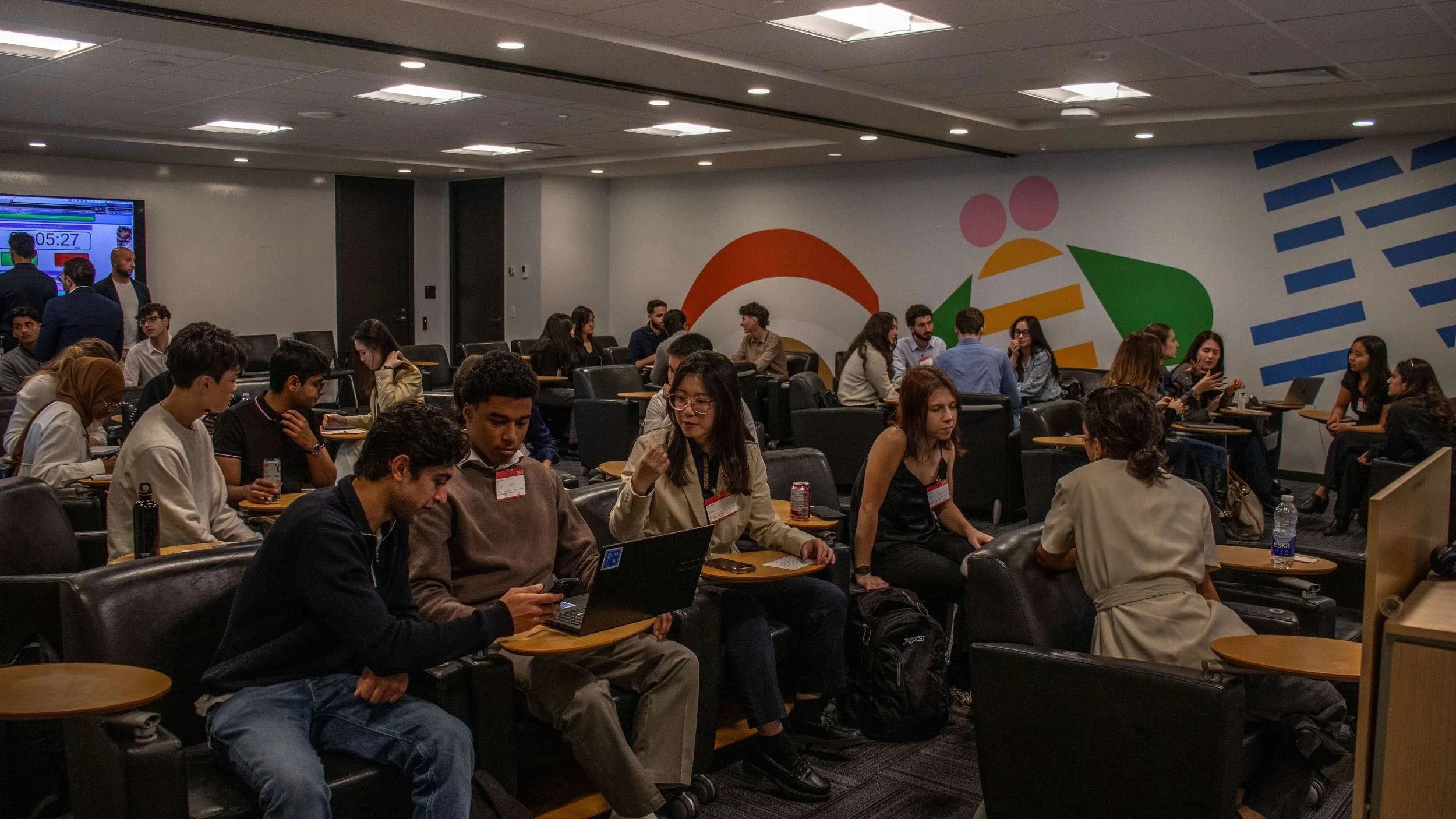 People sitting and talking in a conference room with colorful abstract wall art, some using laptops and smartphones.