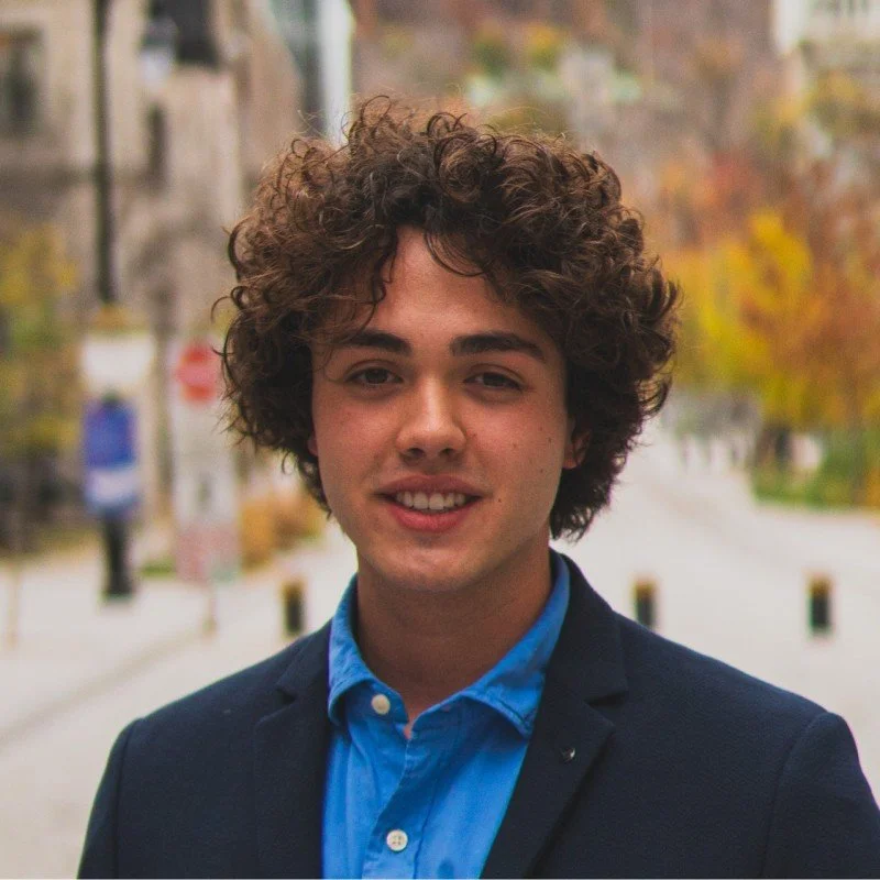 A young man with curly brown hair and light skin smiling outdoors on a city street with autumn trees in the background.