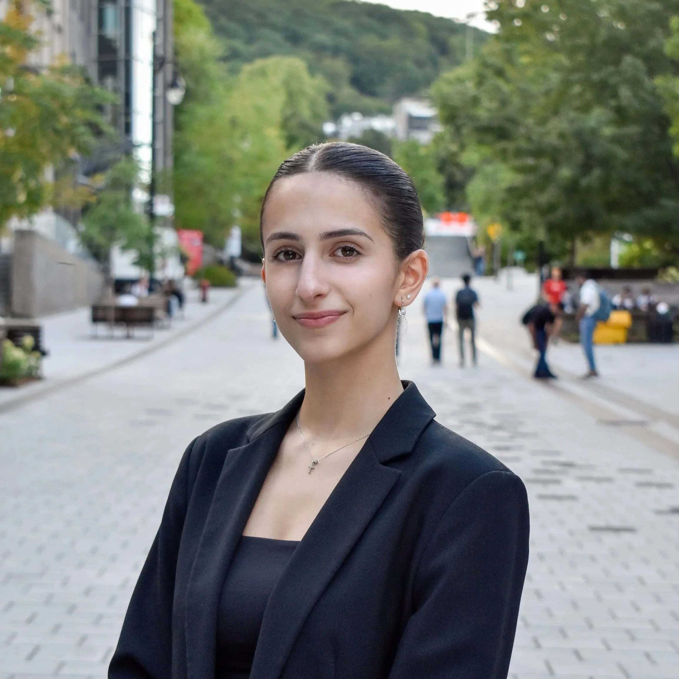 A young woman with dark hair in a bun, wearing a black blazer and a necklace with a cross pendant, standing outdoors on a city street with greenery and people in the background.