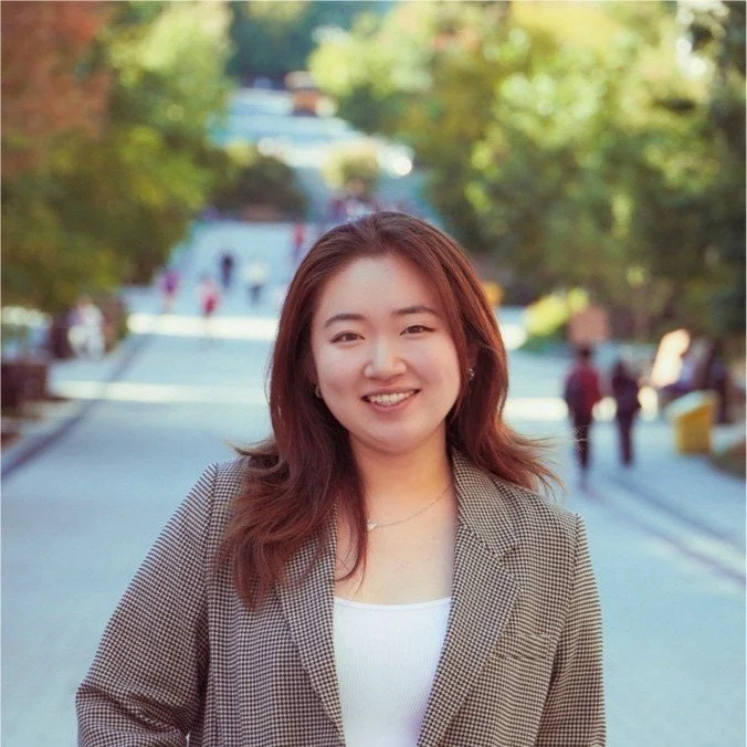 A young woman with shoulder-length brown hair, smiling, standing outdoors on a sunny day with a blurred background of trees and people walking.