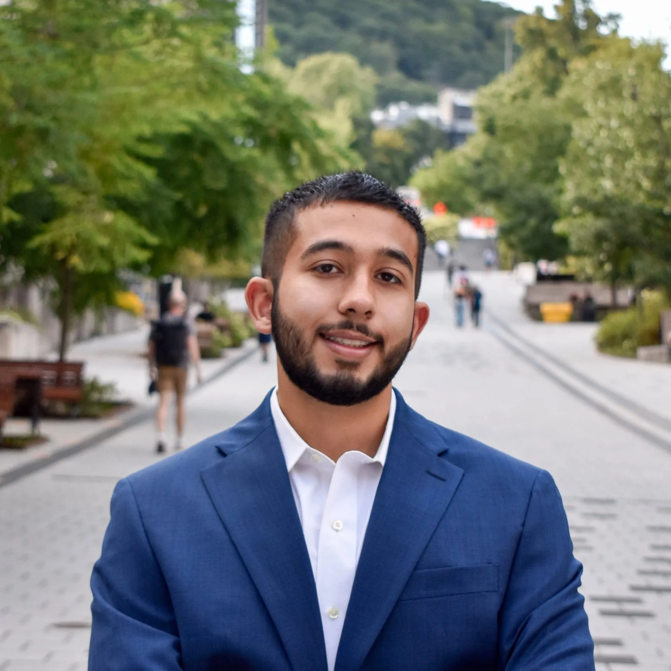A young man with a beard, wearing a blue suit and white shirt, smiling outdoors with a pathway, trees, and people in the background.