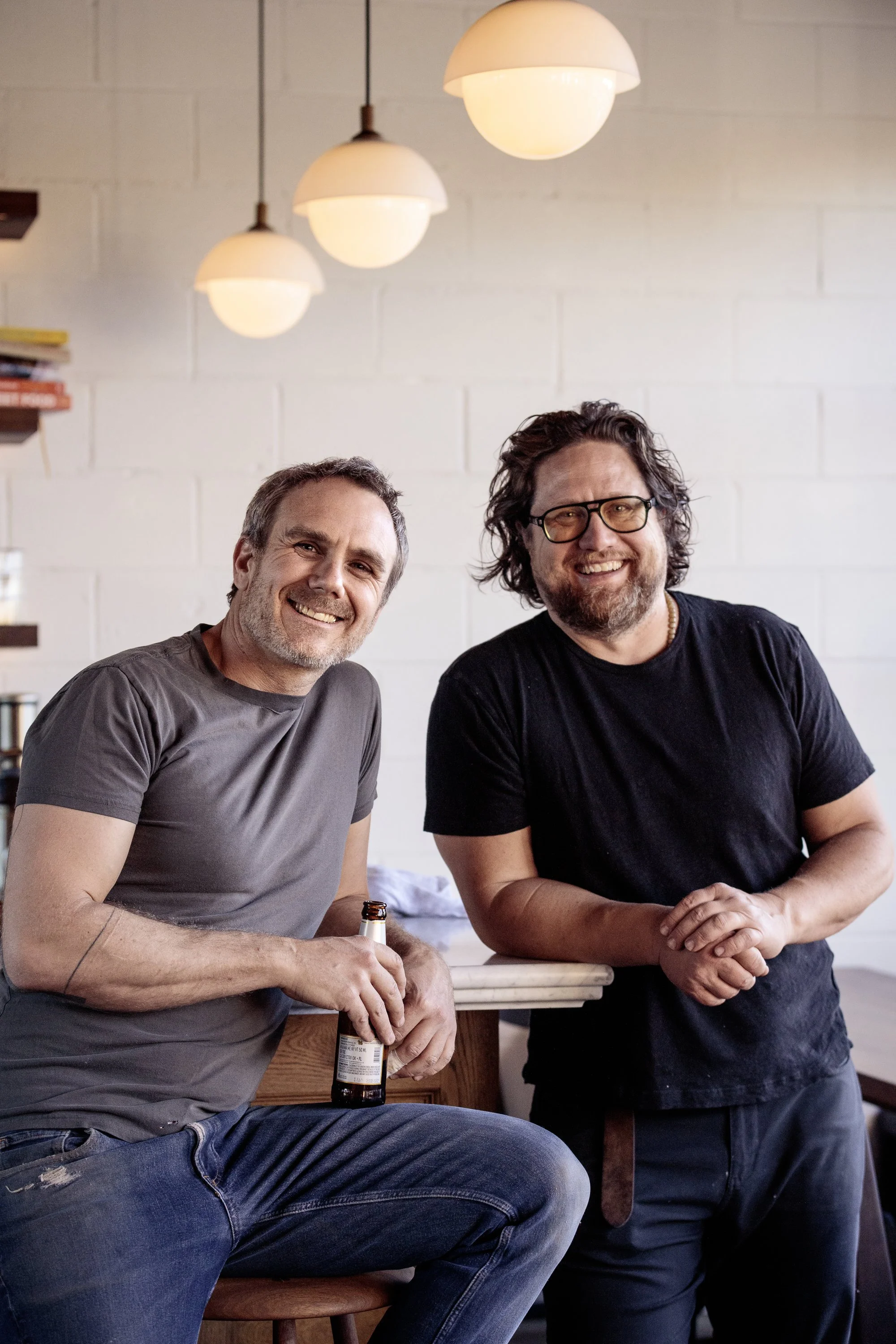 Two smiling men with beards and glasses, standing in a modern, cozy room with white brick walls and hanging pendant lights. One man holds a beer bottle.