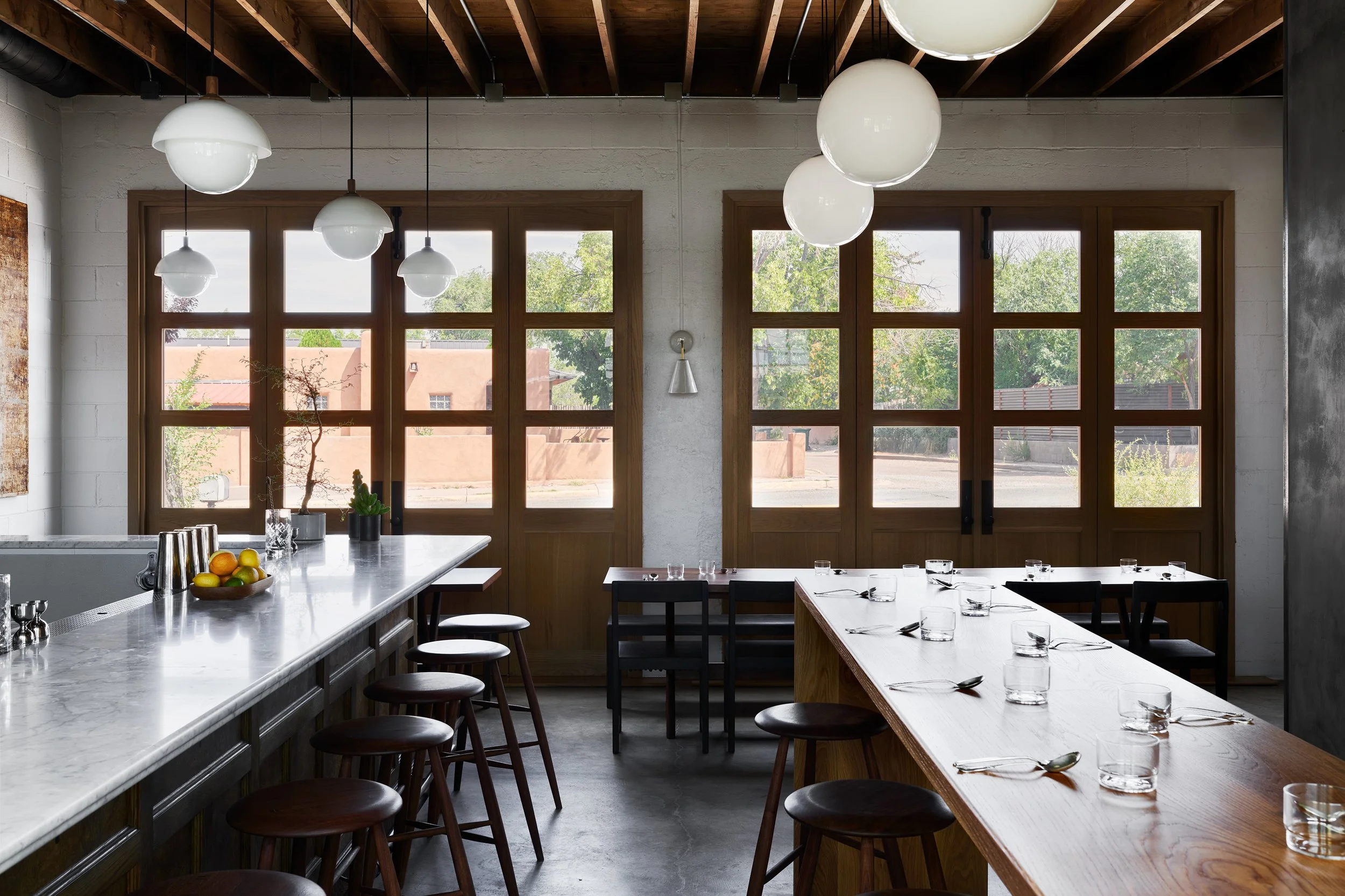 A modern dining area with large windows, pendant lights, a marble countertop with fruit and plants, and a wooden table set with glasses and utensils.