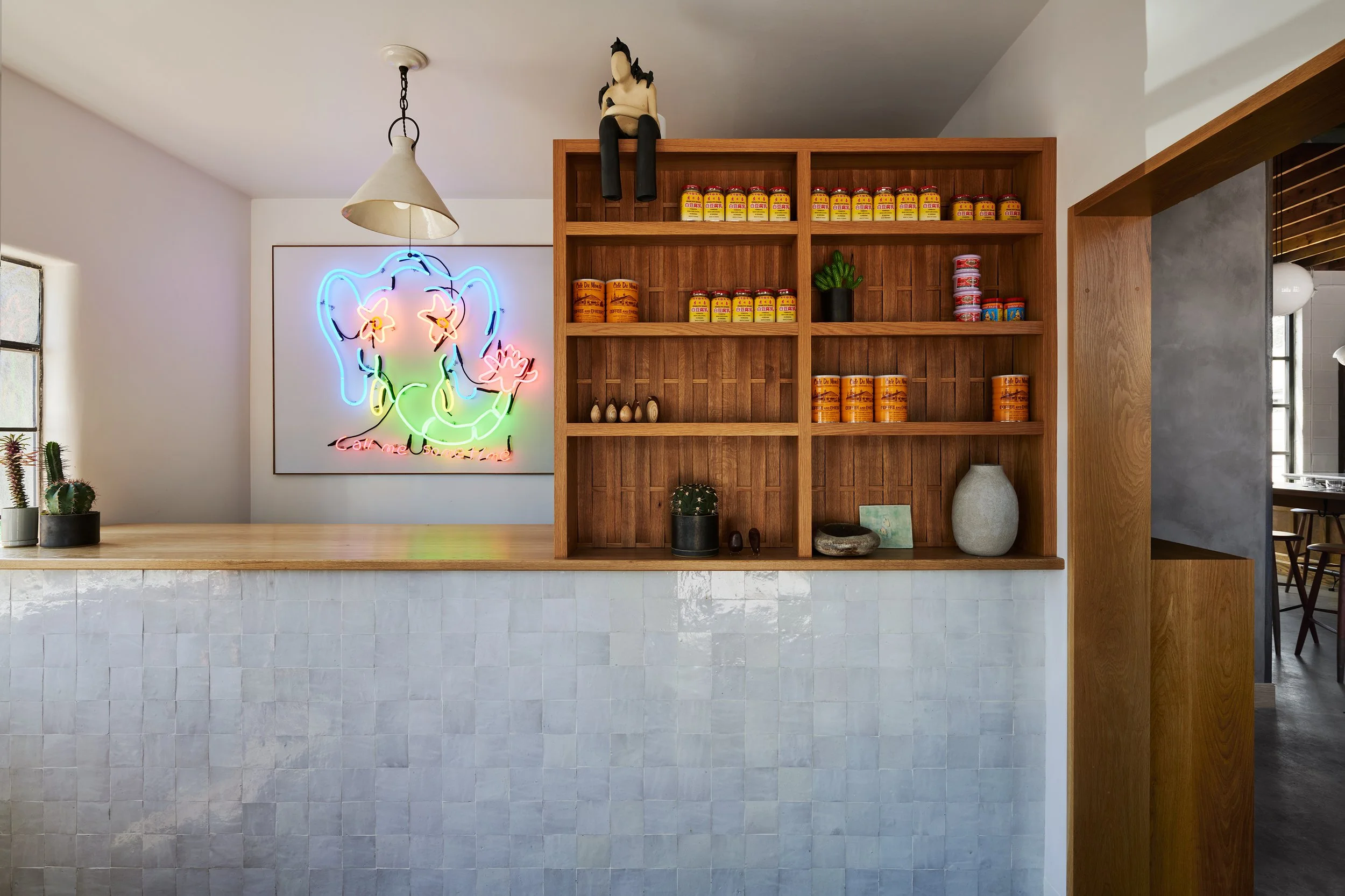 Interior of a cafe with a neon elephant art on the wall, wooden shelves holding canned items, and small decorative objects.